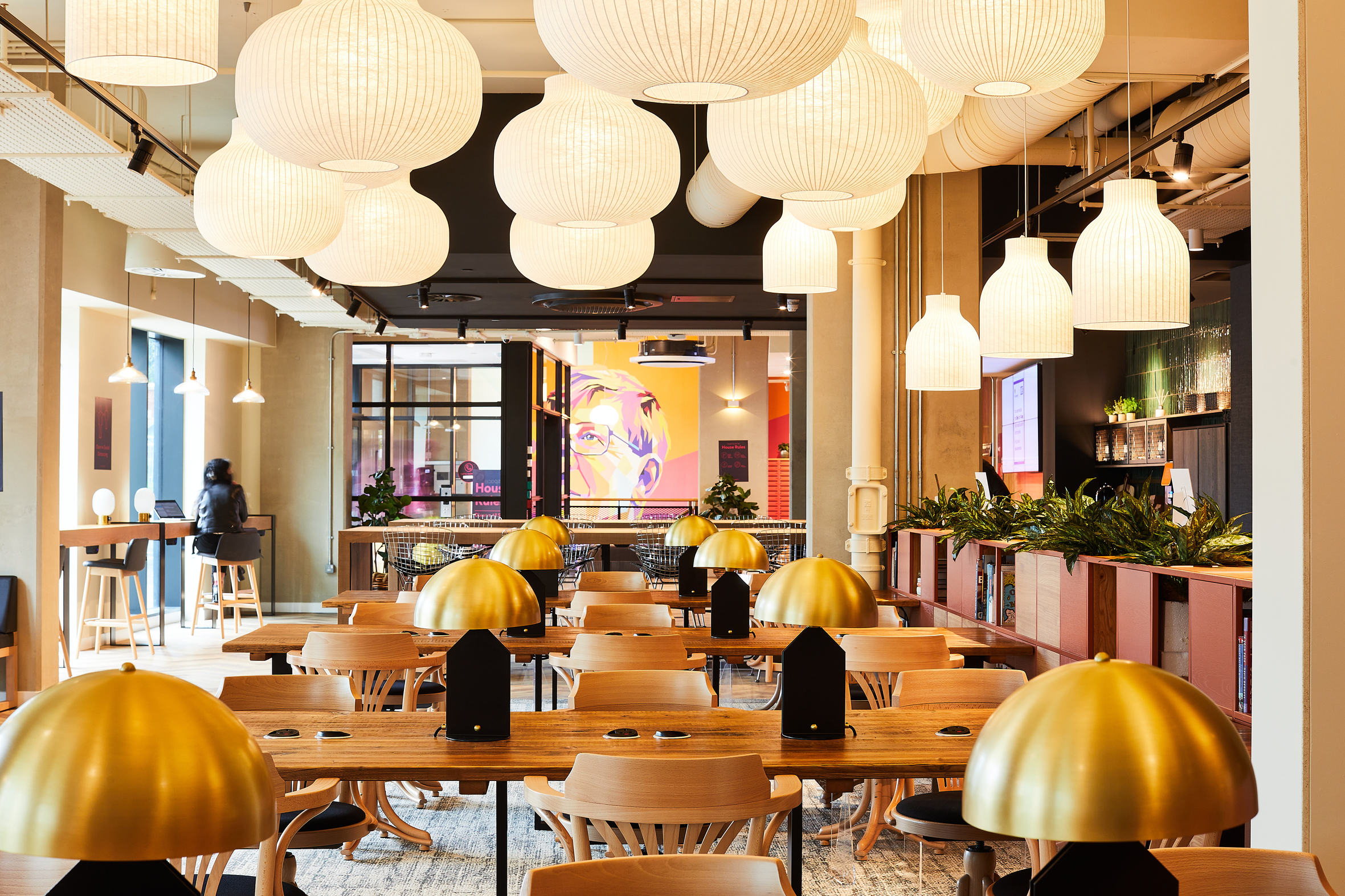 A modern, spacious cafe on Portland Crescent features wooden tables and chairs. Large paper lanterns and gold dome lights hang from the ceiling. Green plants line the walls, and a vivid portrait is visible in the background. A person sits at a high table.