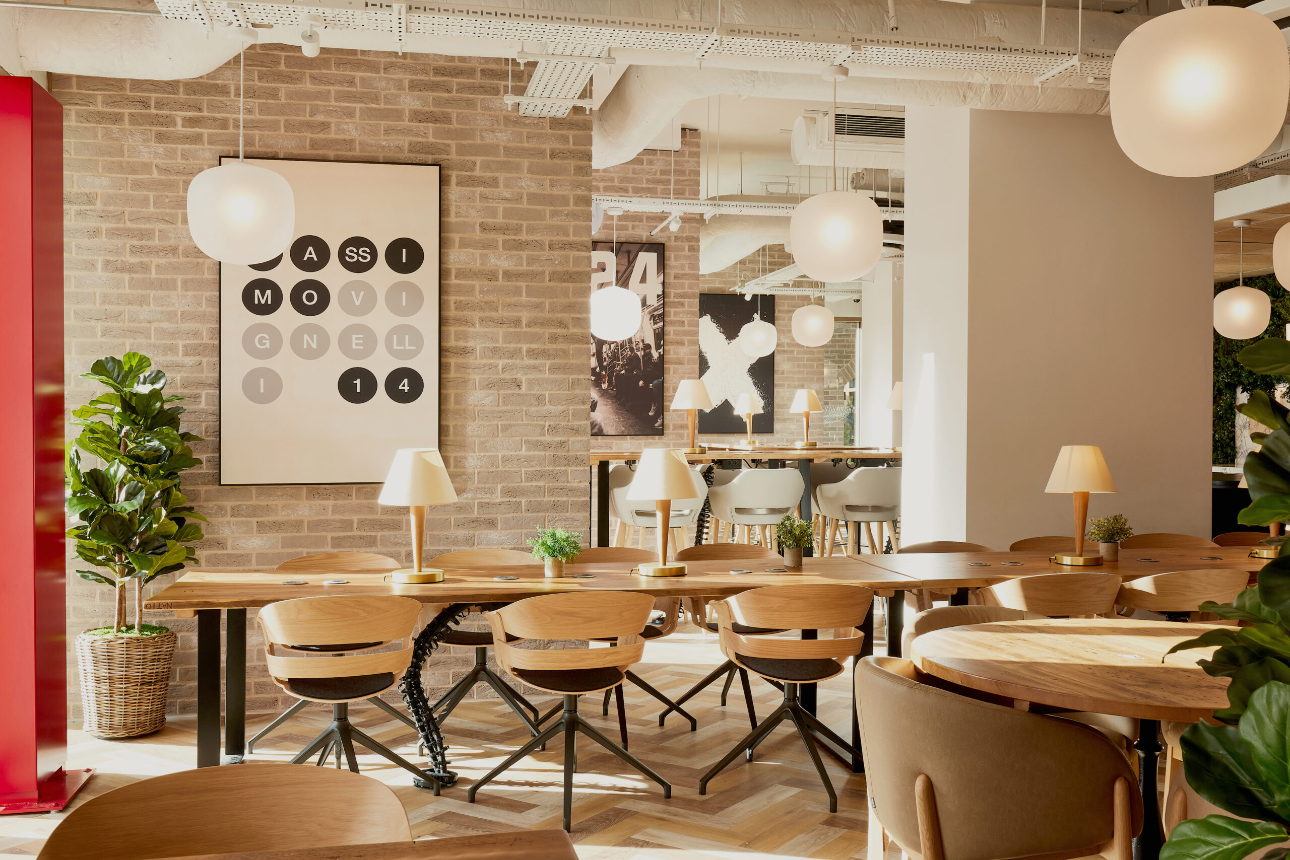 Modern cafe interior with wooden tables and chairs, pendant lights, and a brick wall featuring a large black and white artwork. Potted plants add greenery, and mirrors enhance the cozy, inviting atmosphere.