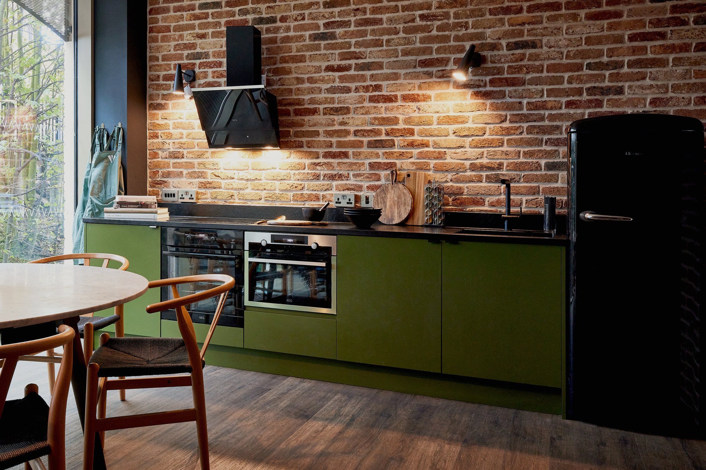 A modern kitchen in the Lewisham Exchange, boasting green cabinets and brick walls. It features a black refrigerator, built-in oven, and stove. Wooden cutting boards and utensils adorn the counter. A round table with chairs sits in the foreground, while a large window lets in natural light.