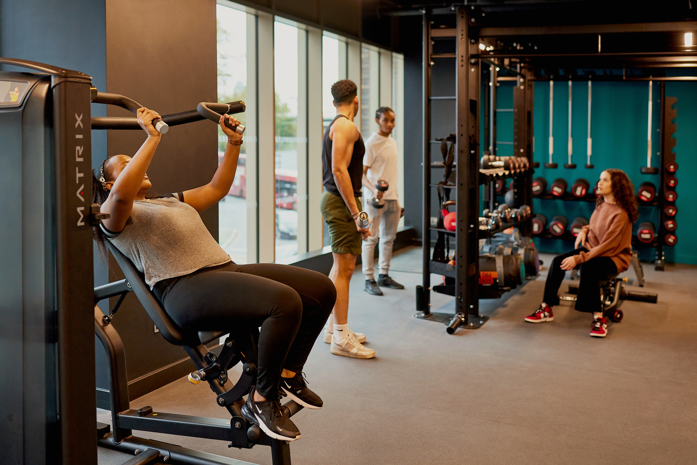 A woman exercises on a machine at the spacious Lewisham Exchange gym, while two men and a woman chat, holding weights in the background. The gym's large windows flood the area with natural light, highlighting the array of equipment available.