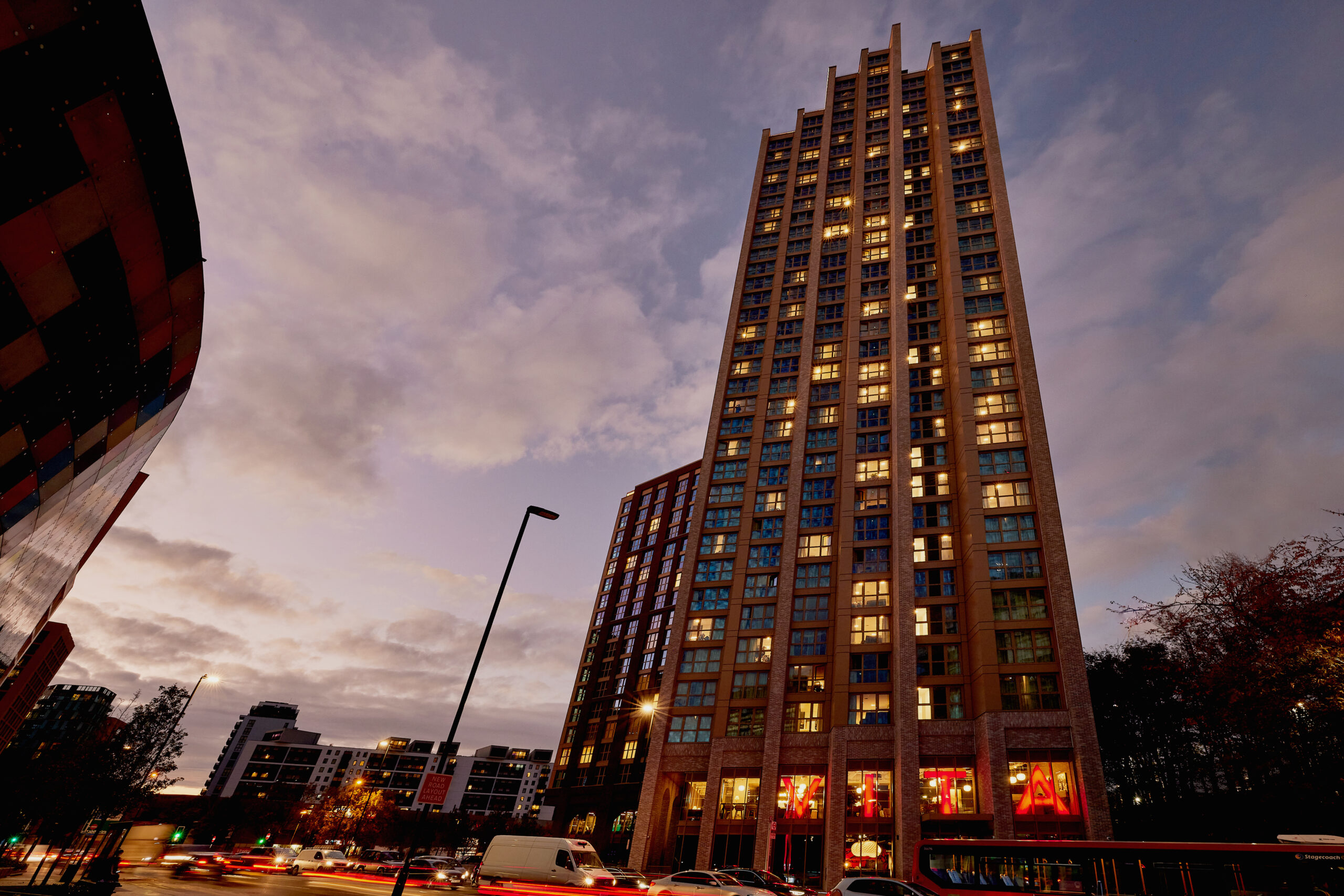 Tall building illuminated at dusk stands against a cloudy sky. Its windows glow warmly, while trees and a curved building flank it. Streetlights and blurred traffic lights below suggest a busy urban setting.