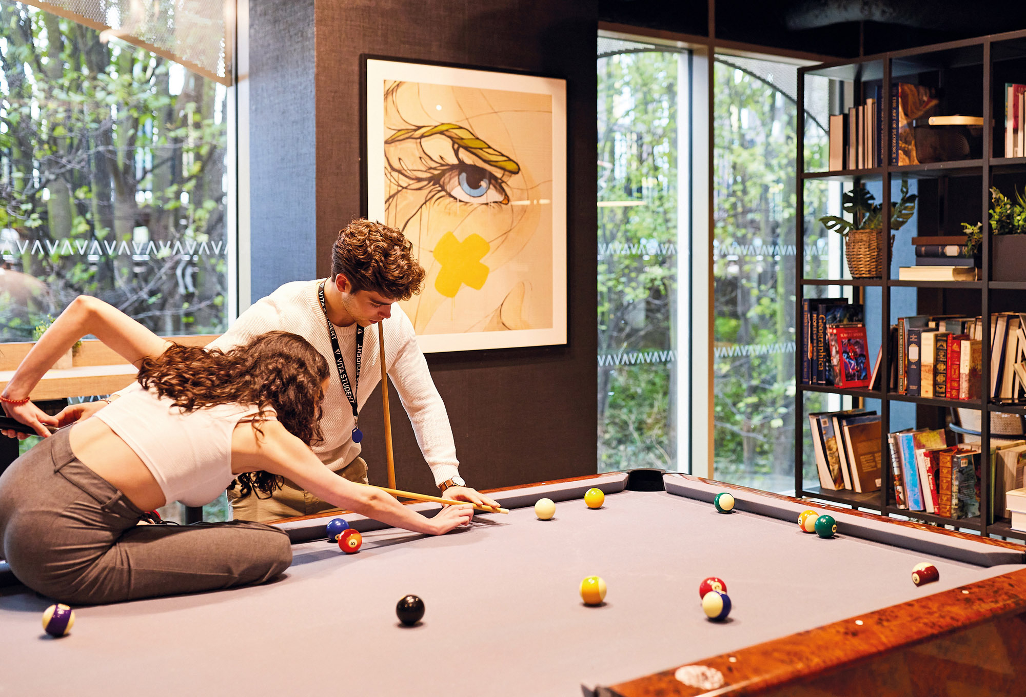 In a cozy room with a large window, a woman and man engage in a lively exchange over billiards. The woman leans in, aiming her cue as the man assists. Behind them, vibrant artwork adds flair, while a bookshelf brims with books and decor.