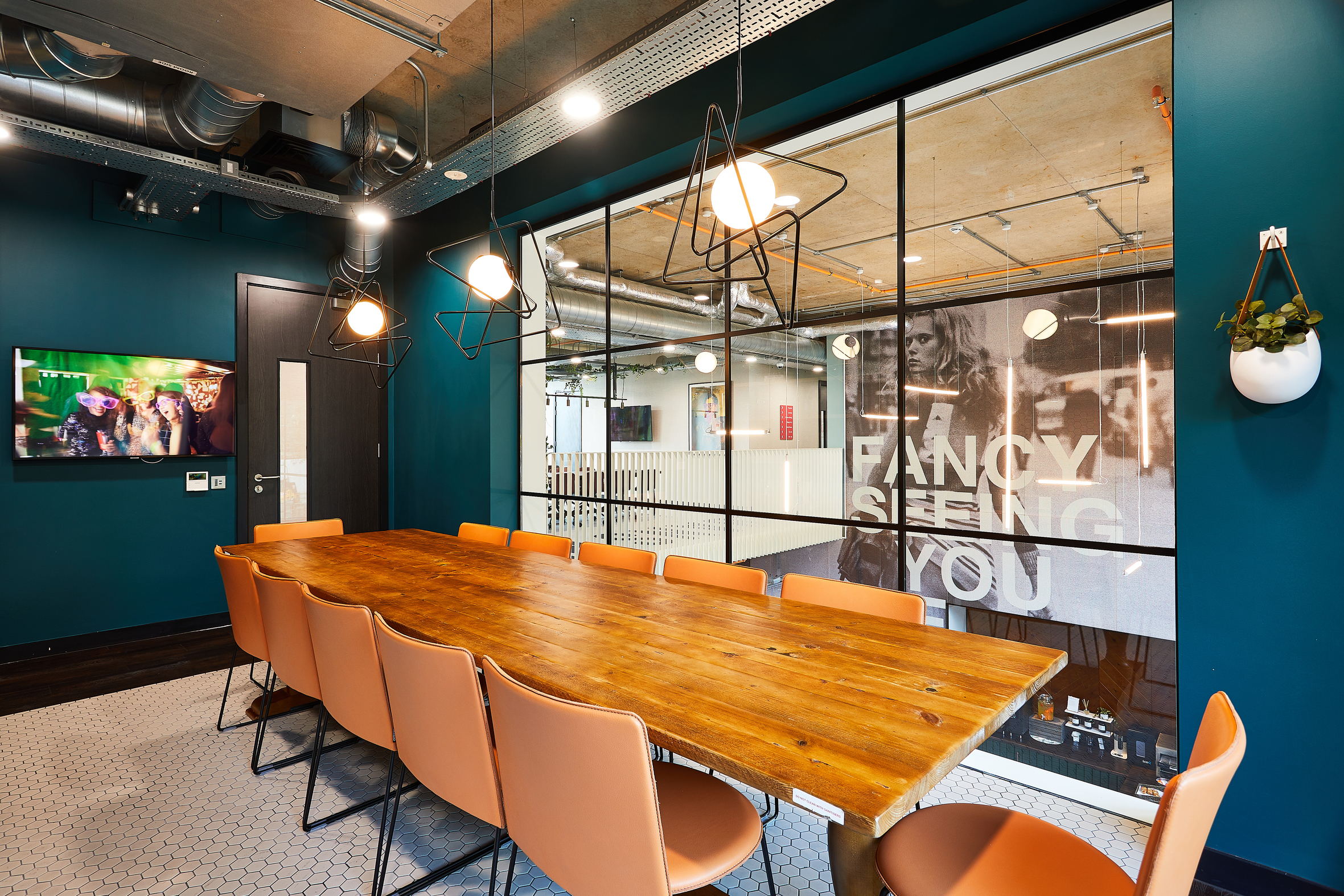 A modern meeting room with a wooden table surrounded by eight tan chairs. The walls are teal, and a large window displays the phrase "FANCY SEEING YOU." Industrial lighting and exposed ducts add to the contemporary design.