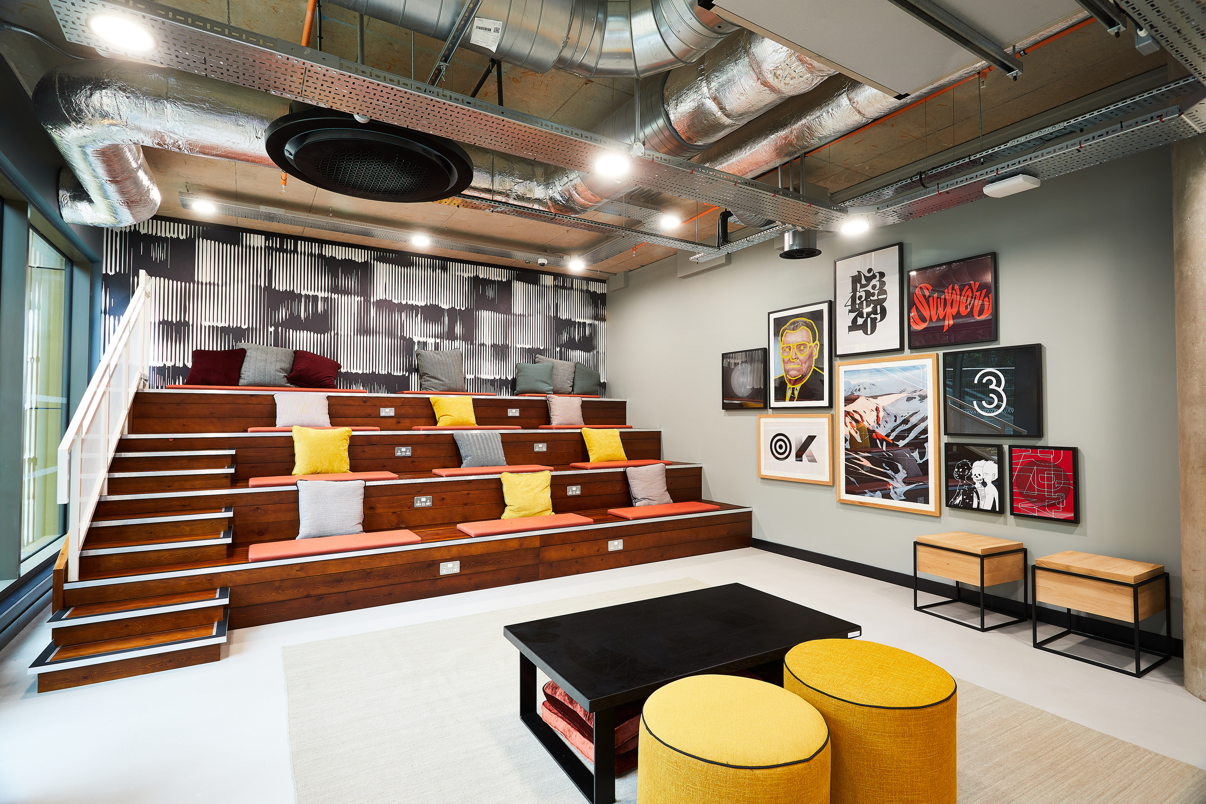 Modern room with tiered wooden seating, brightly colored cushions, and a variety of framed art on the walls. Exposed ductwork on the ceiling, stools, a black coffee table, and round yellow ottomans complete the eclectic decor.