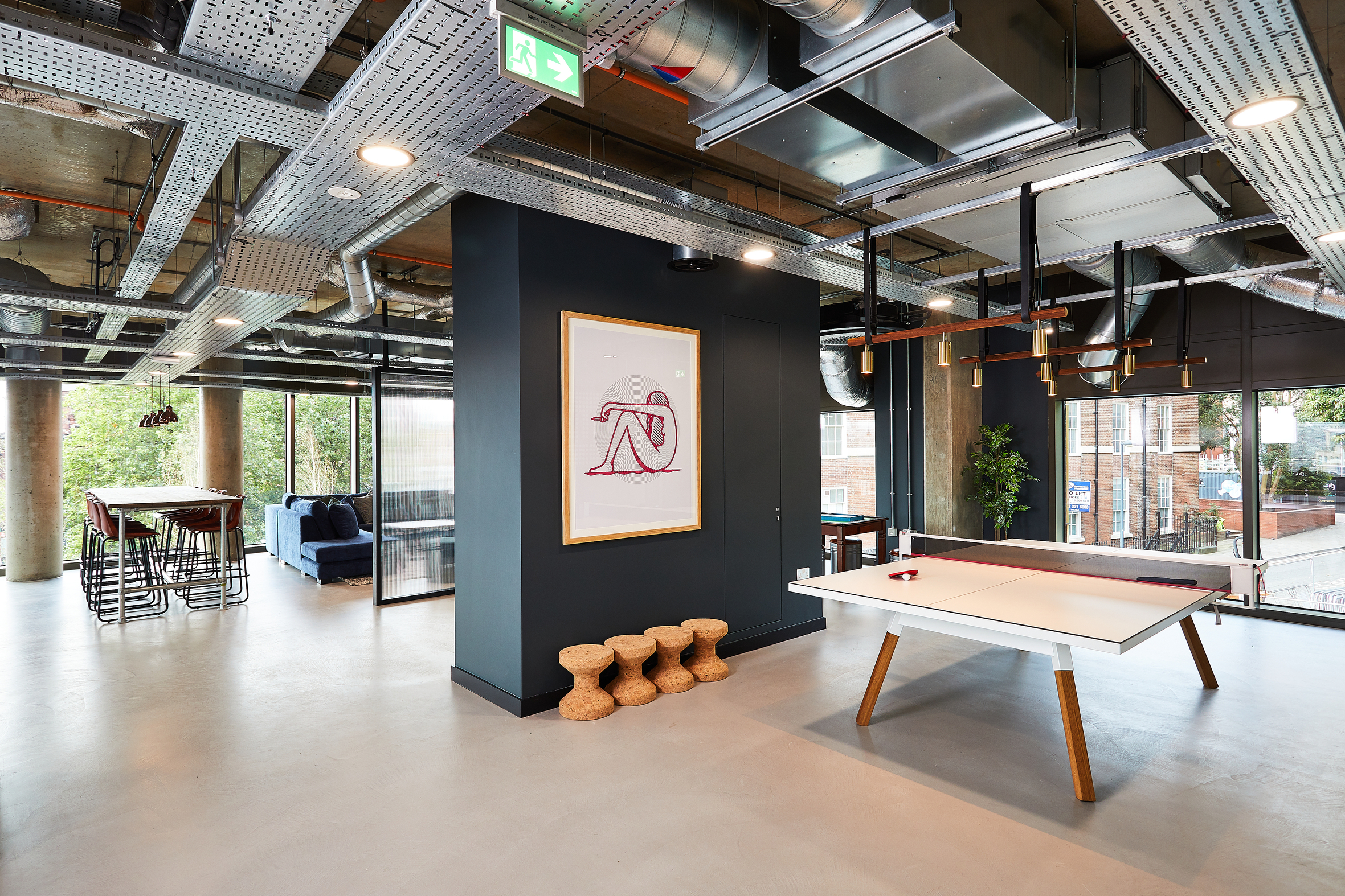 Modern office space with industrial design features a white ping pong table, a large framed art piece on a dark wall, and a seating area with a long table and high chairs. Exposed ducts and large windows add to the spacious feel.