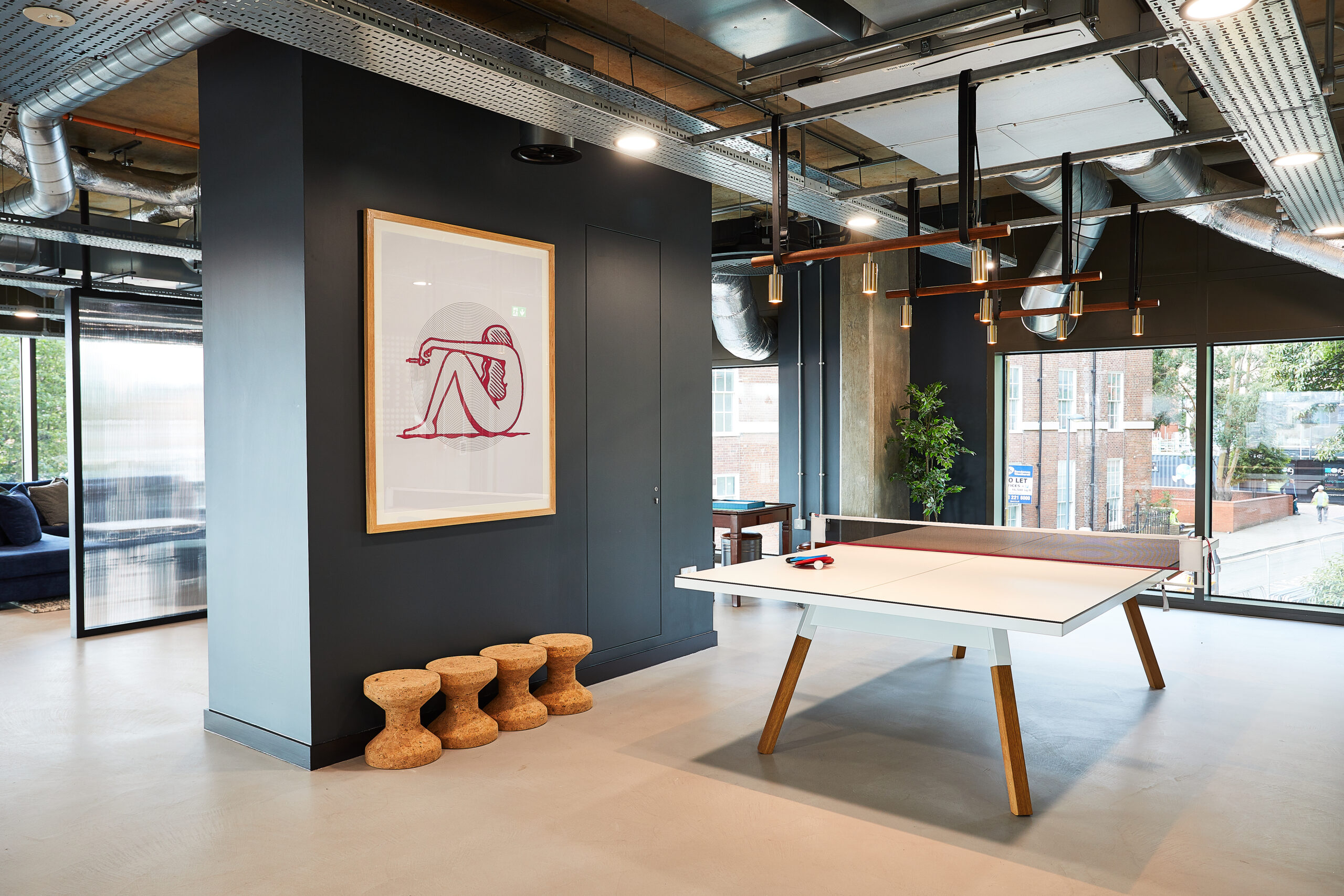 Modern room with a ping pong table and paddles on it. A large window shows an urban view. There's a framed artwork on the wall and three wooden stools nearby. Exposed ductwork and ambient lighting add to the contemporary design.