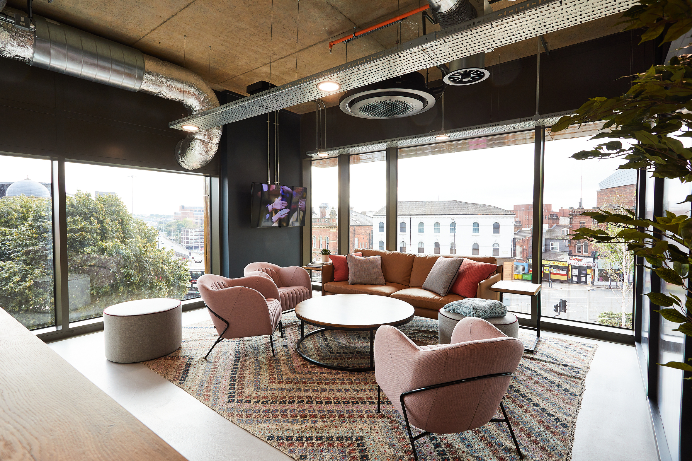 Modern office lounge with large windows, featuring a brown sofa, four pink armchairs, and a round coffee table on a patterned rug. There's visible ductwork on the ceiling and a wall-mounted TV. Outdoor buildings and trees are seen through the windows.