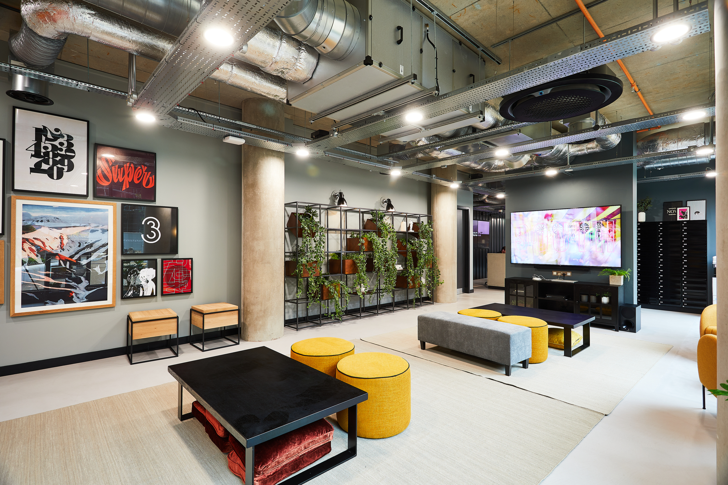 Modern lounge area with industrial design, featuring exposed pipes and concrete columns. Decor includes framed art, a large TV, and plants. Seating includes black and yellow poufs, benches, and a grey couch on a light rug.
