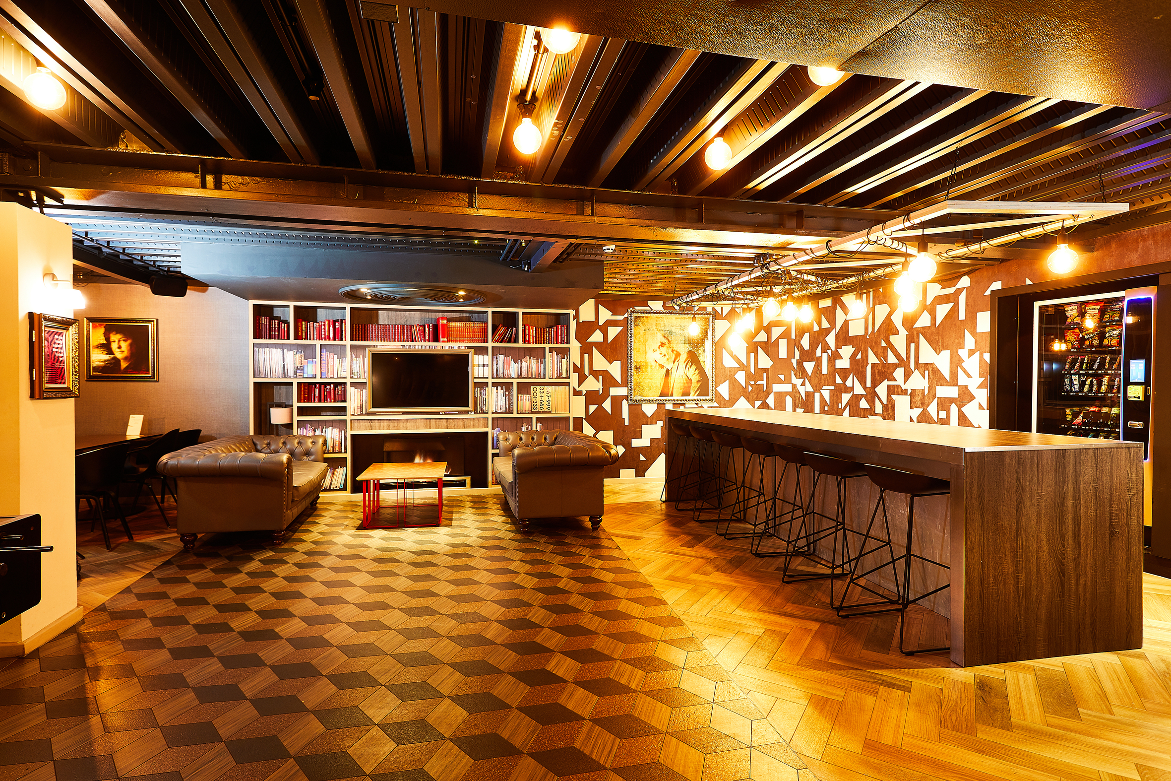 This luxury student accommodation boasts a modern, warmly lit lounge with geometric-patterned floors and walls. It features bookshelves, a TV, two grey sofas, and a long bar with stools. Art decorates the space, complemented by lights hanging from black ceiling beams and vending machines on the right.