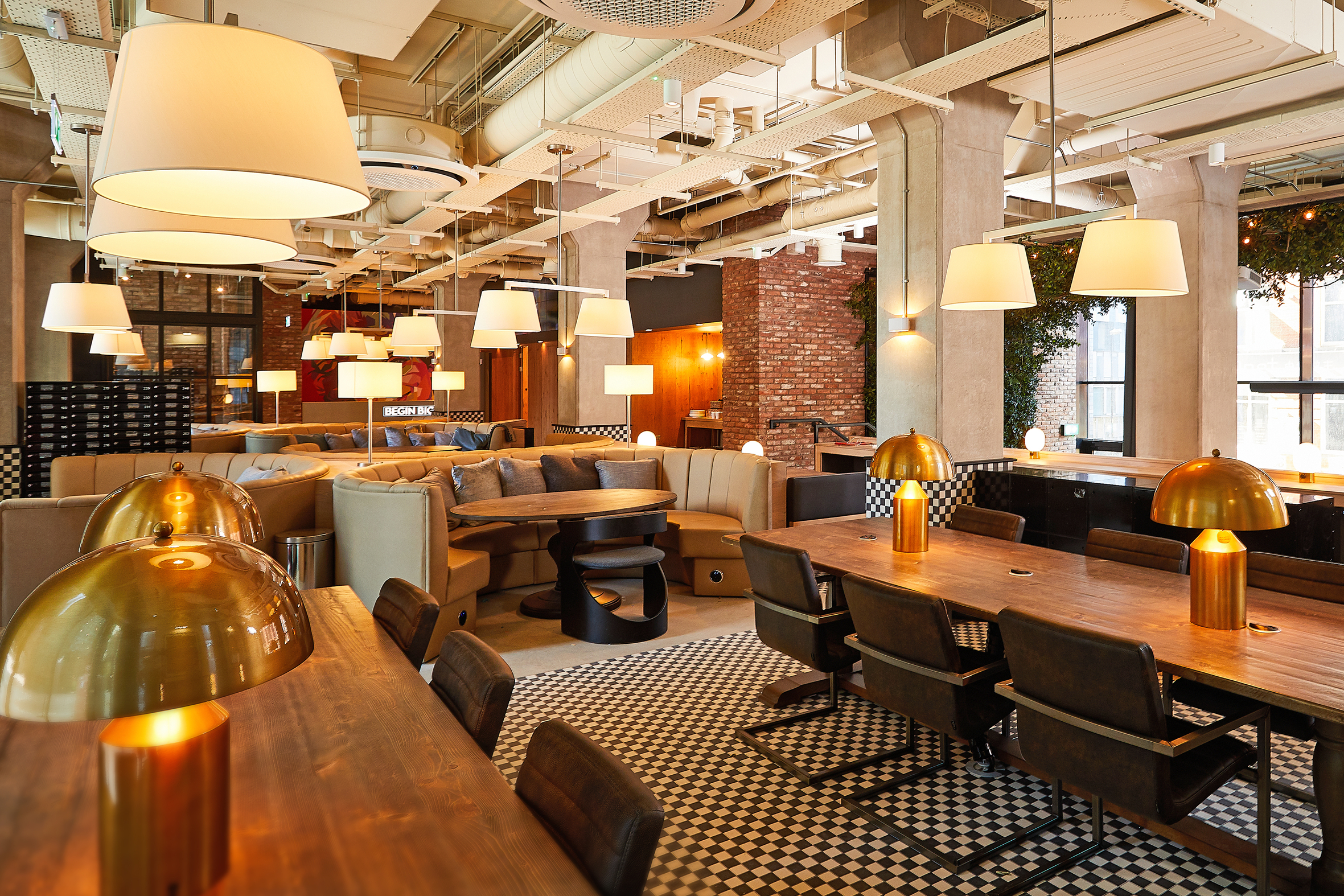 Modern restaurant interior with warm lighting, featuring wooden tables, square brown chairs, and gold dome lamps. The space has a checkered floor, brick walls, and large circular windows. Cozy booth seating lines the perimeter, and hanging lights create a welcoming atmosphere.