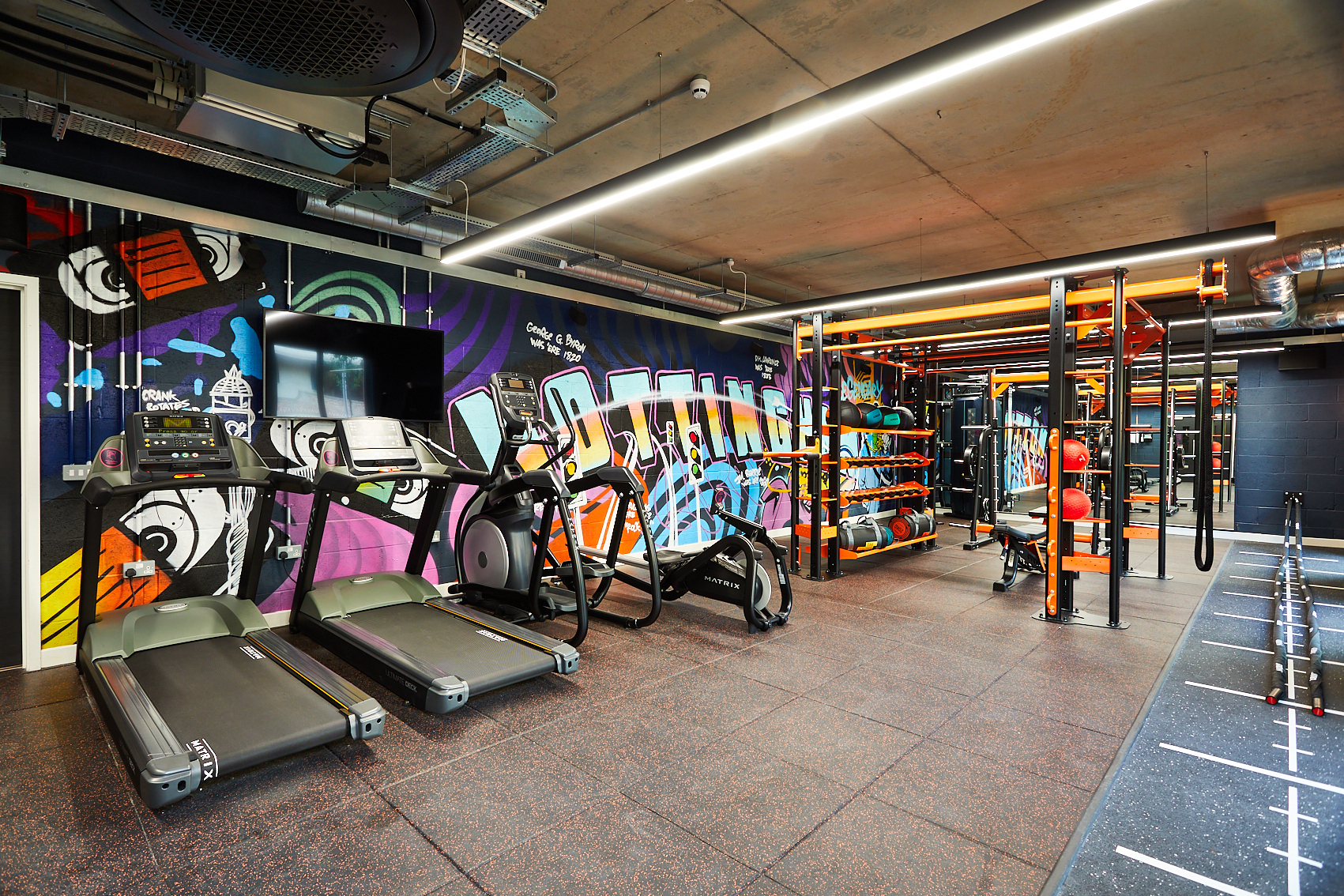 Located on Station Street, this modern gym features treadmills, exercise bikes, and a weight rack in a functional training area. A colorful graffiti mural graces one wall, while bright lighting accentuates the state-of-the-art equipment.