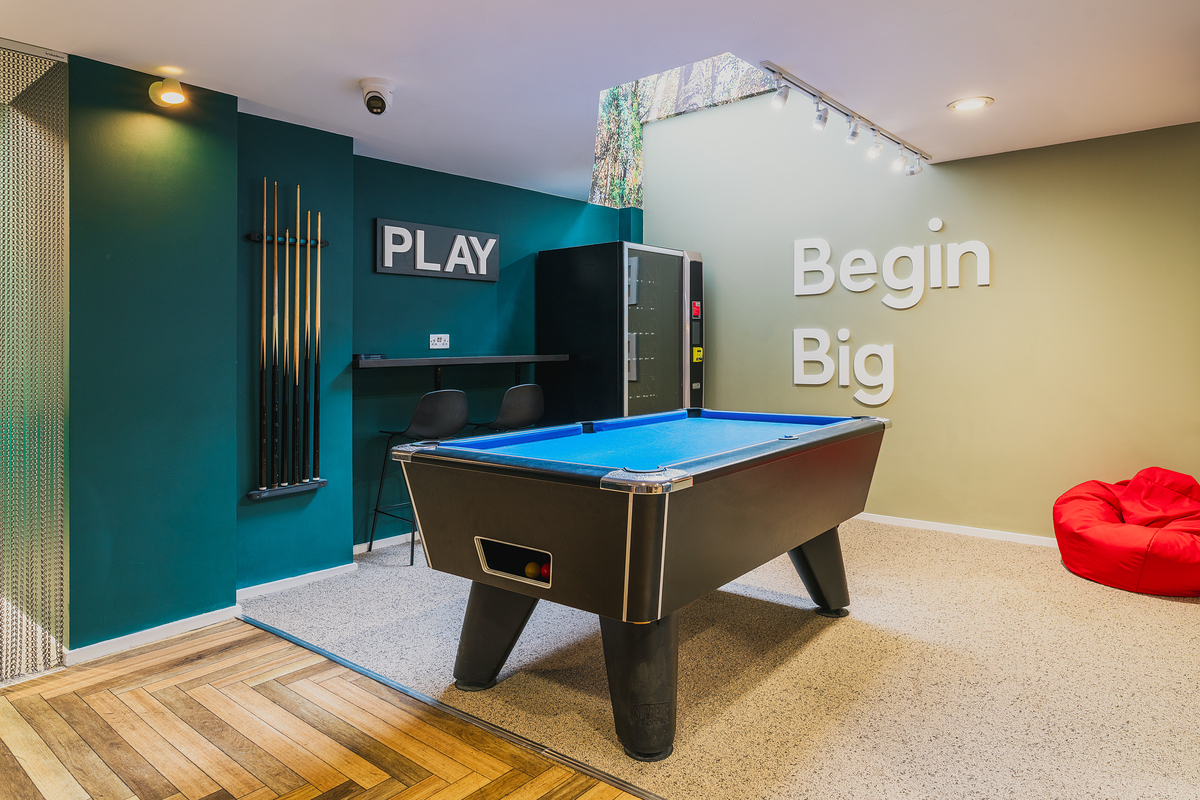 A modern game room at Portland House features a blue pool table, two black chairs, a wall-mounted cue rack, a vending machine, a red bean bag, and wall text reading "PLAY" and "Begin Big" under skylight lighting.