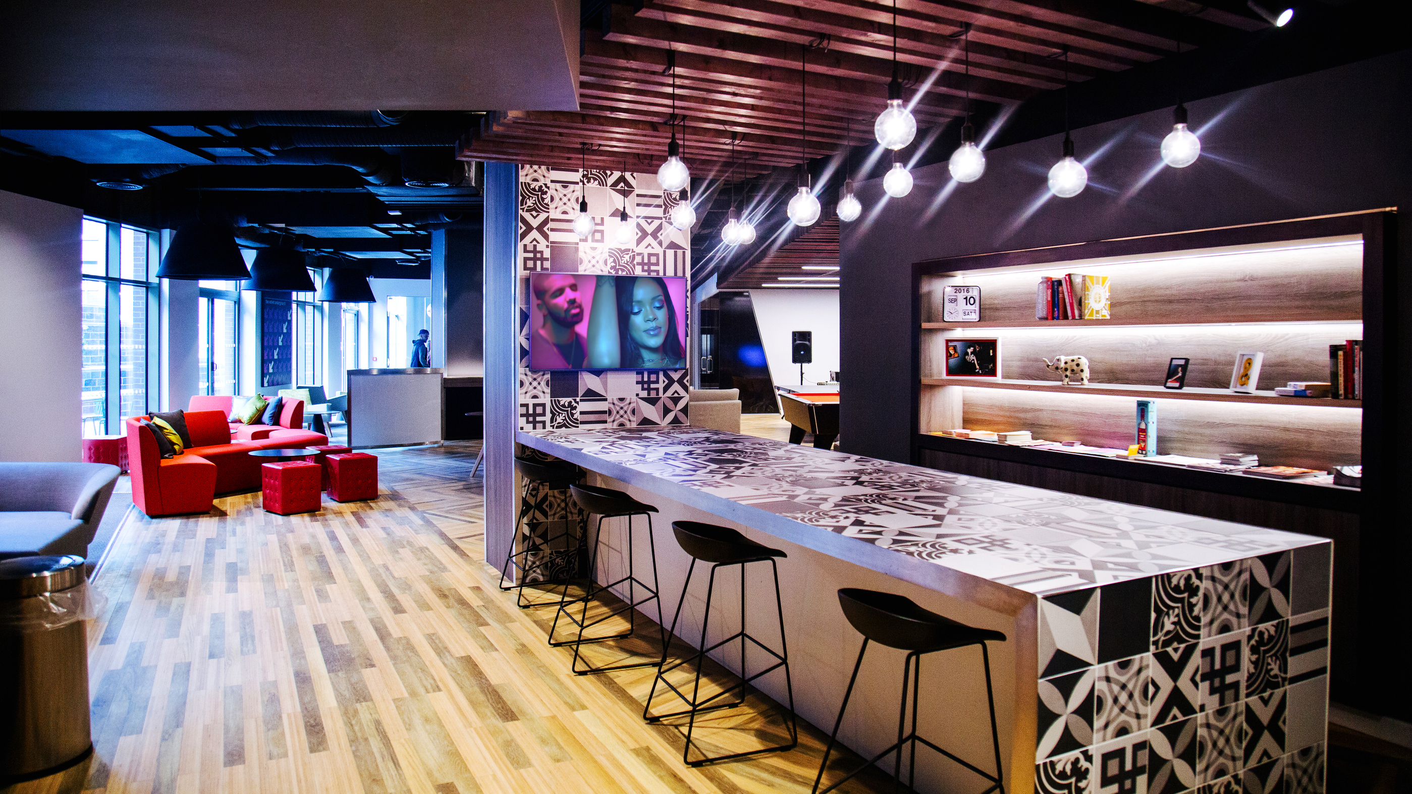 Modern office lounge with a patterned bar counter, five bar stools, and hanging lights. Shelves with books and decor items line one wall. Red chairs and a sofa are in the seating area by large windows, allowing natural light in.