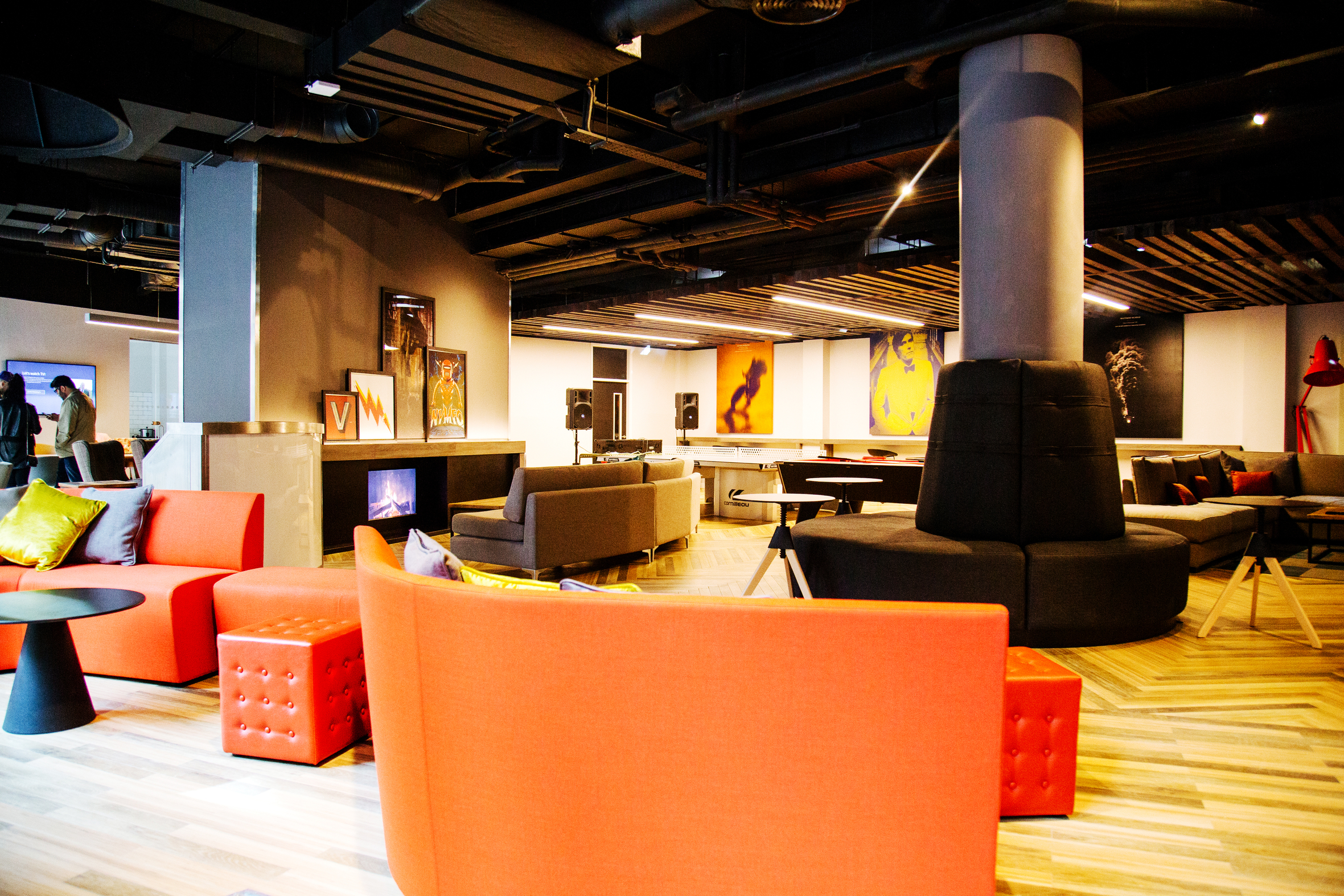 A modern lounge area featuring vibrant red and orange seating, including couches and cushioned stools. The space has a cozy fireplace, contemporary artwork, and a mix of wooden and dark ceilings, creating a stylish and inviting atmosphere.