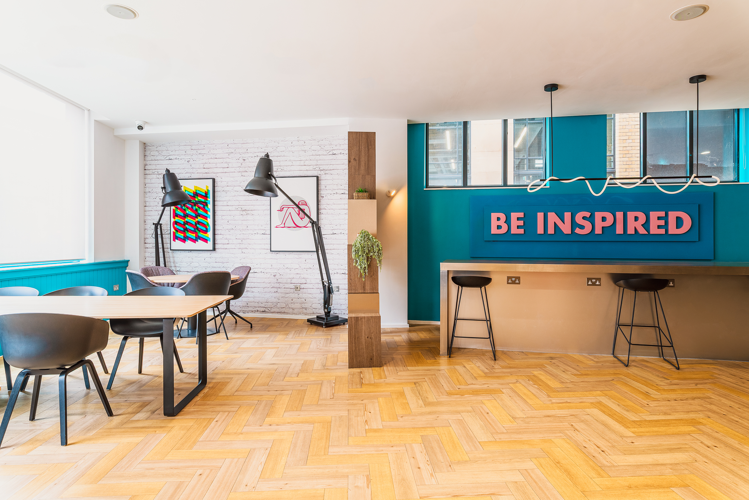 A bright, modern workspace with wooden chevron flooring, tables and chairs, artwork on a white brick wall, large black lamps, and a teal accent wall displaying "BE INSPIRED" in bold pink letters.