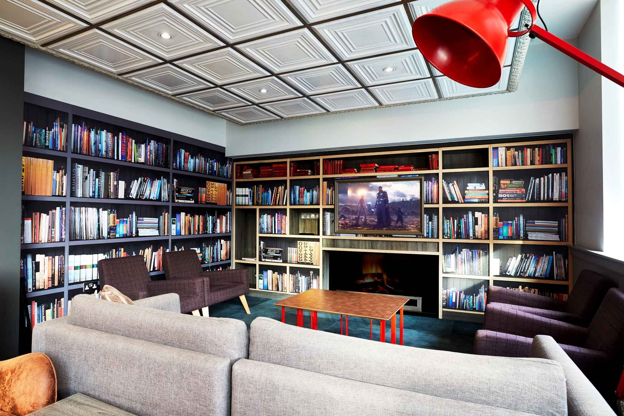 A cozy library with black bookshelves filled with books surrounds a seating area. A large TV above a modern fireplace displays a movie. There are gray sofas and armchairs, and a red floor lamp stands in the corner. The ceiling is white with recessed lights.