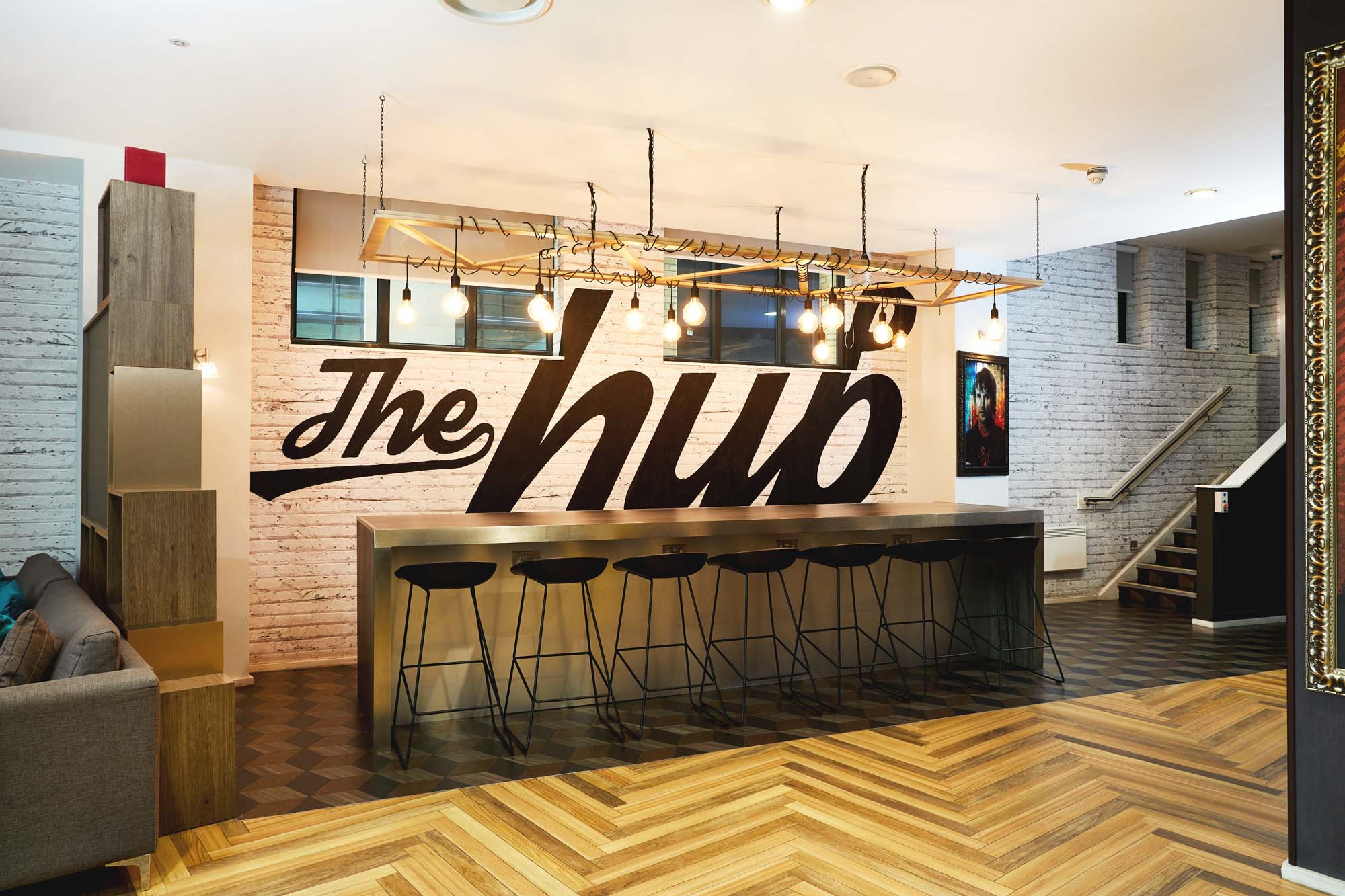 Modern interior space with a long bar counter and high stools, beneath a decorative light fixture. The wall features the words "The Hub" in large black script. The floor has a parquet pattern, and stairs lead to an upper level.