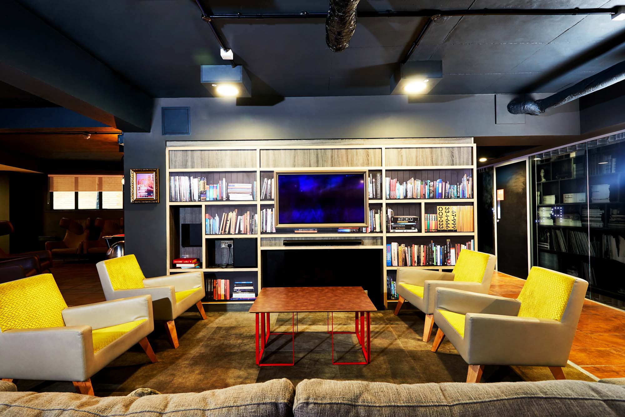 A modern lounge area with four yellow armchairs surrounding a square coffee table. Behind them, a large bookshelf holds books and a flat-screen TV. The ceiling and walls are dark, and the space has a contemporary, cozy atmosphere.