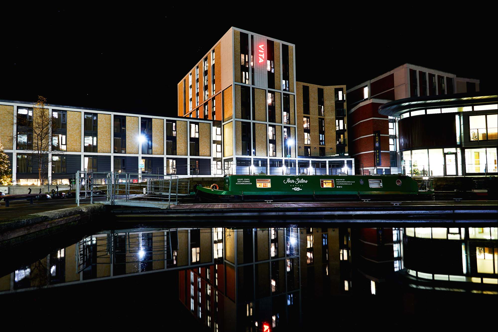 Night view of a modern multi-story building complex illuminated with lights, reflecting in a calm canal. A green narrowboat is docked in the water, and the structure is adorned with various rectangular windows and a sign with "VIA" on it.