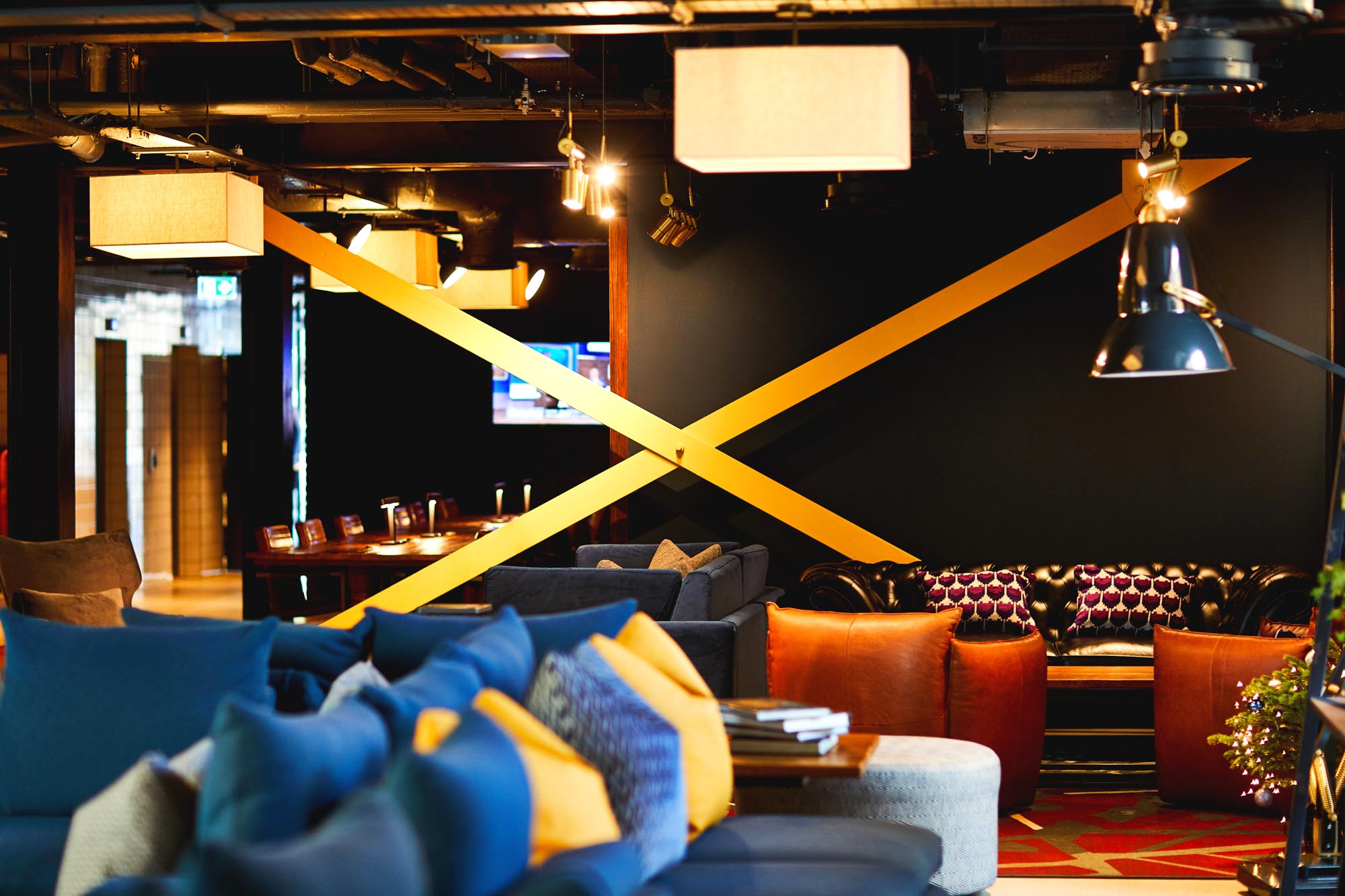 A cozy lounge area with a mix of blue and orange sofas and cushions. The room features modern lighting, a dark wall with yellow crossed beams, and a leather chair. There is a small table with books, creating a warm and inviting atmosphere.