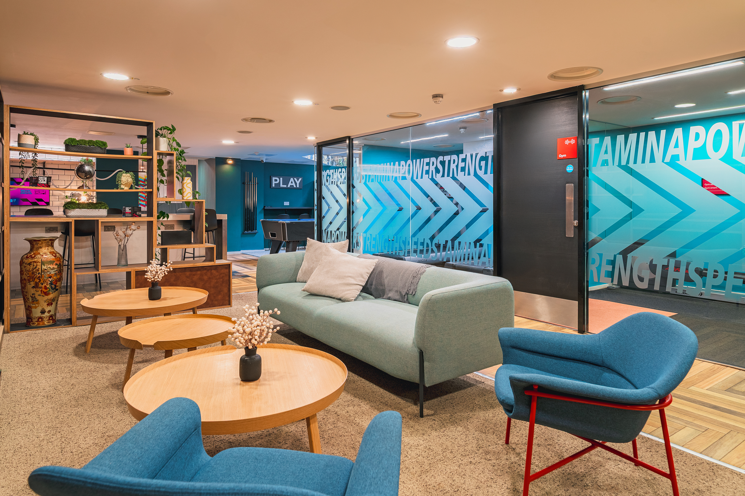 A modern lounge area at Portland House features a light blue sofa, two blue armchairs, and round wooden tables. Decorative shelves, vases, and plants accent the space. Glass walls display bold text patterns in a signature blue theme.