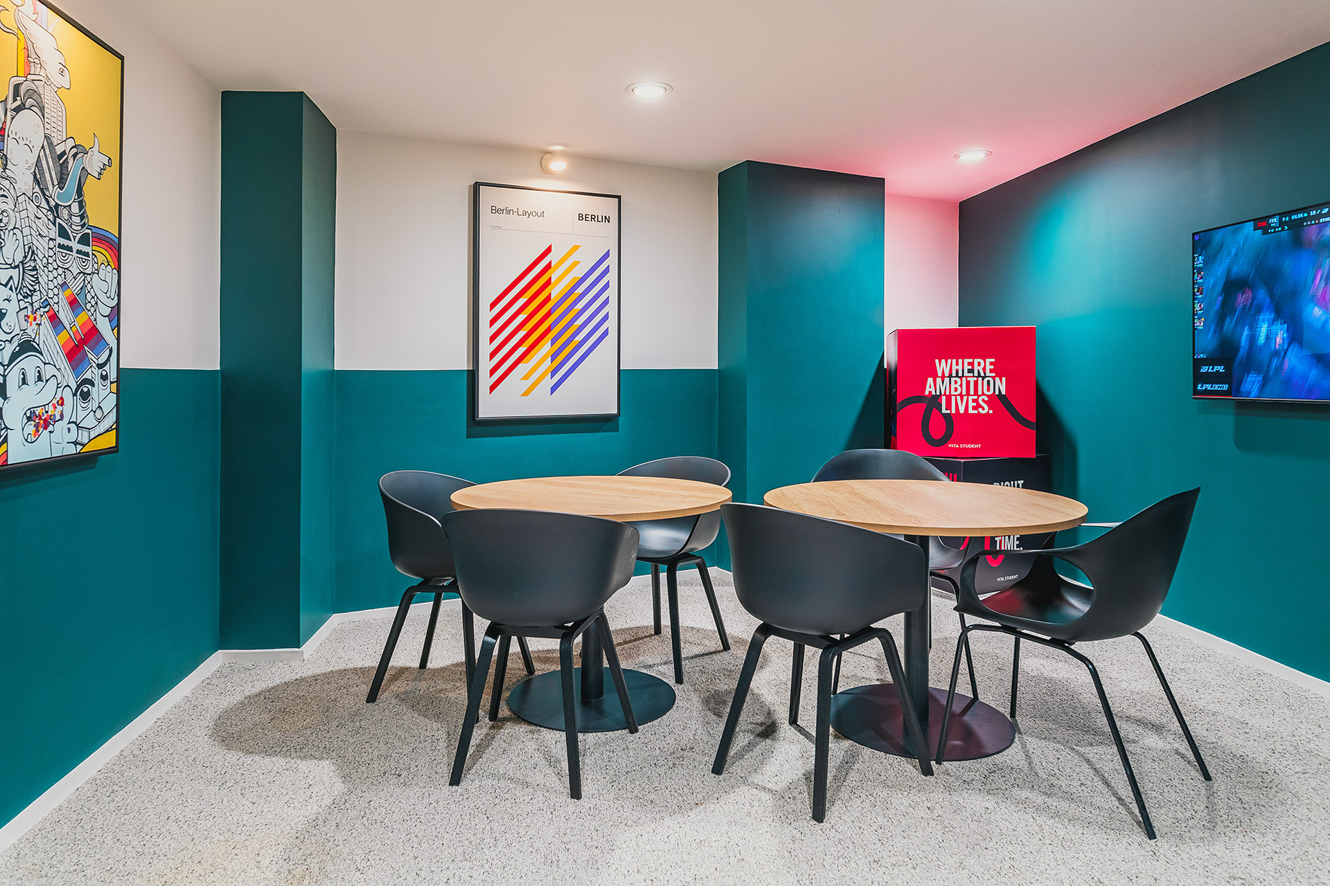A modern meeting room at Portland House features two round wooden tables, six black chairs, colorful abstract art, teal and white walls, a TV screen, and a red sign reading "Where Ambition Lives.