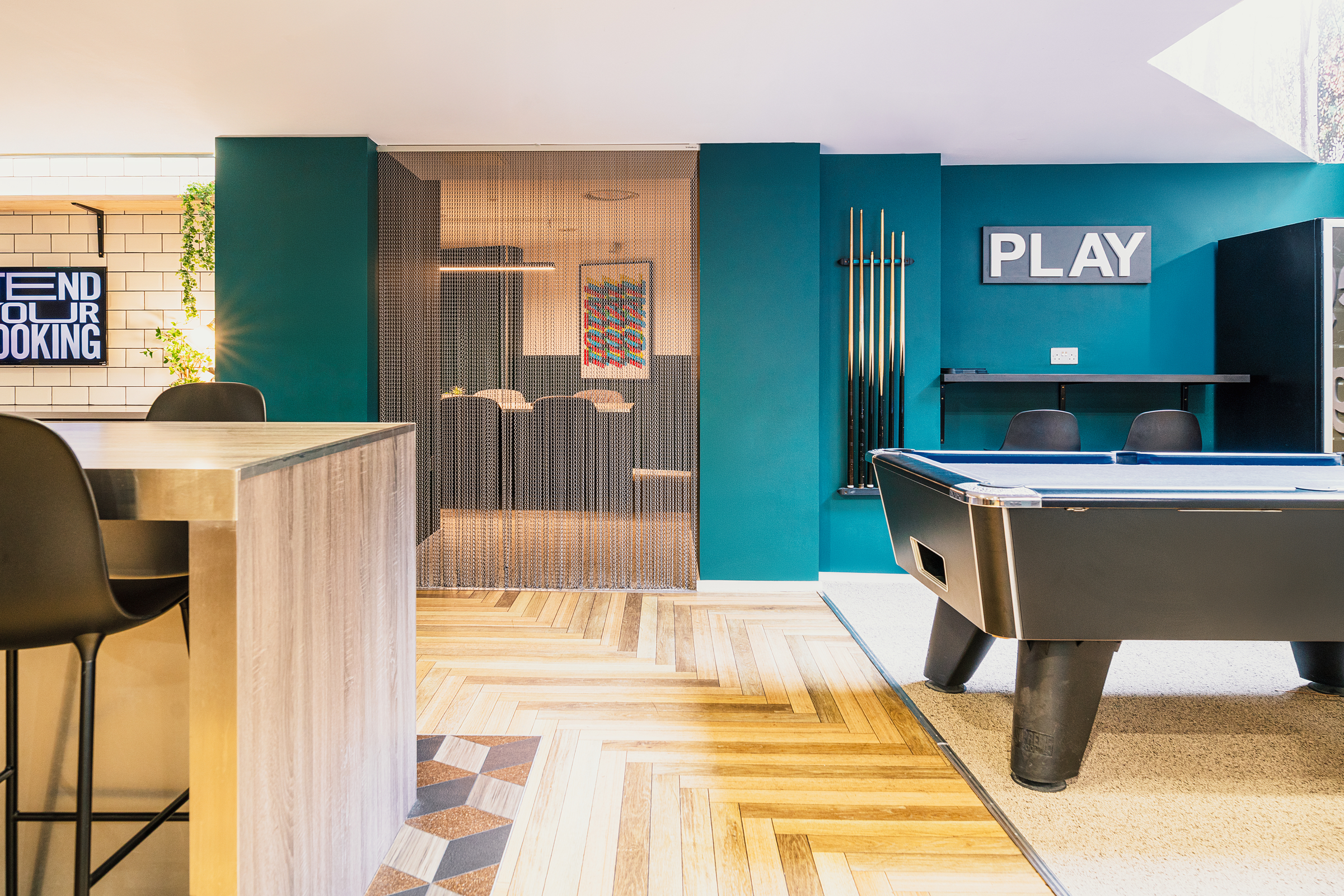 Modern game room in the Portland House with teal walls, a pool table, cues on a rack, tall black chairs at a wooden counter, and a "PLAY" sign on the wall. The geometric wood floor adds stylish flair.
