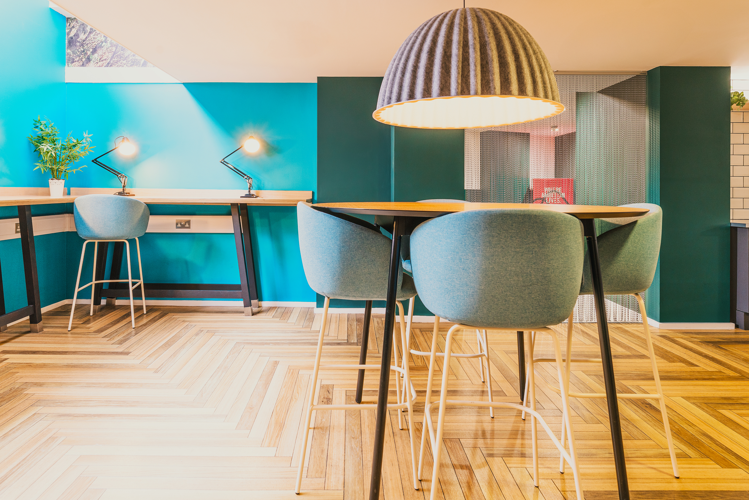 A modern, brightly lit workspace at Portland House with teal walls, blue chairs, wooden tables, a large pendant lamp, and chevron-patterned wooden flooring. Two desks with lamps and small plants sit against the back wall.