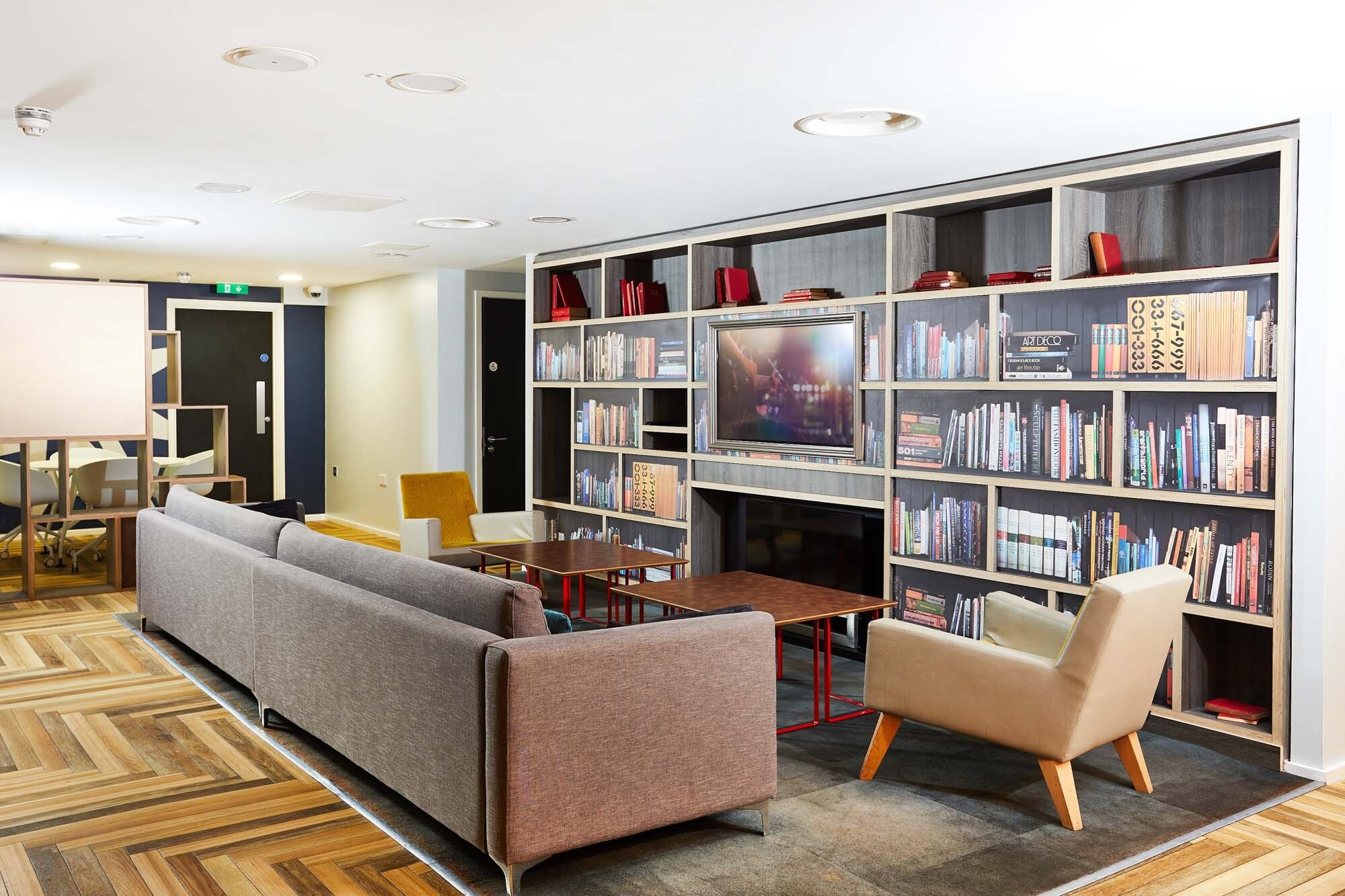 Modern lounge in a Portland House, featuring wood flooring with a gray sofa, two beige chairs, and a wooden coffee table. A large bookshelf filled with books and decorative items graces the back wall, while a TV is mounted at its center.