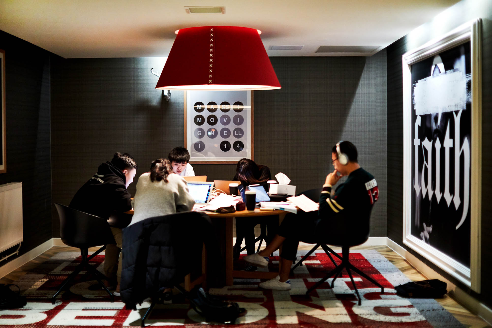 Five people sit around a table in a dimly lit room, working on laptops. A large red lamp hangs above them, and art is displayed on the walls. The setting appears casual and collaborative.