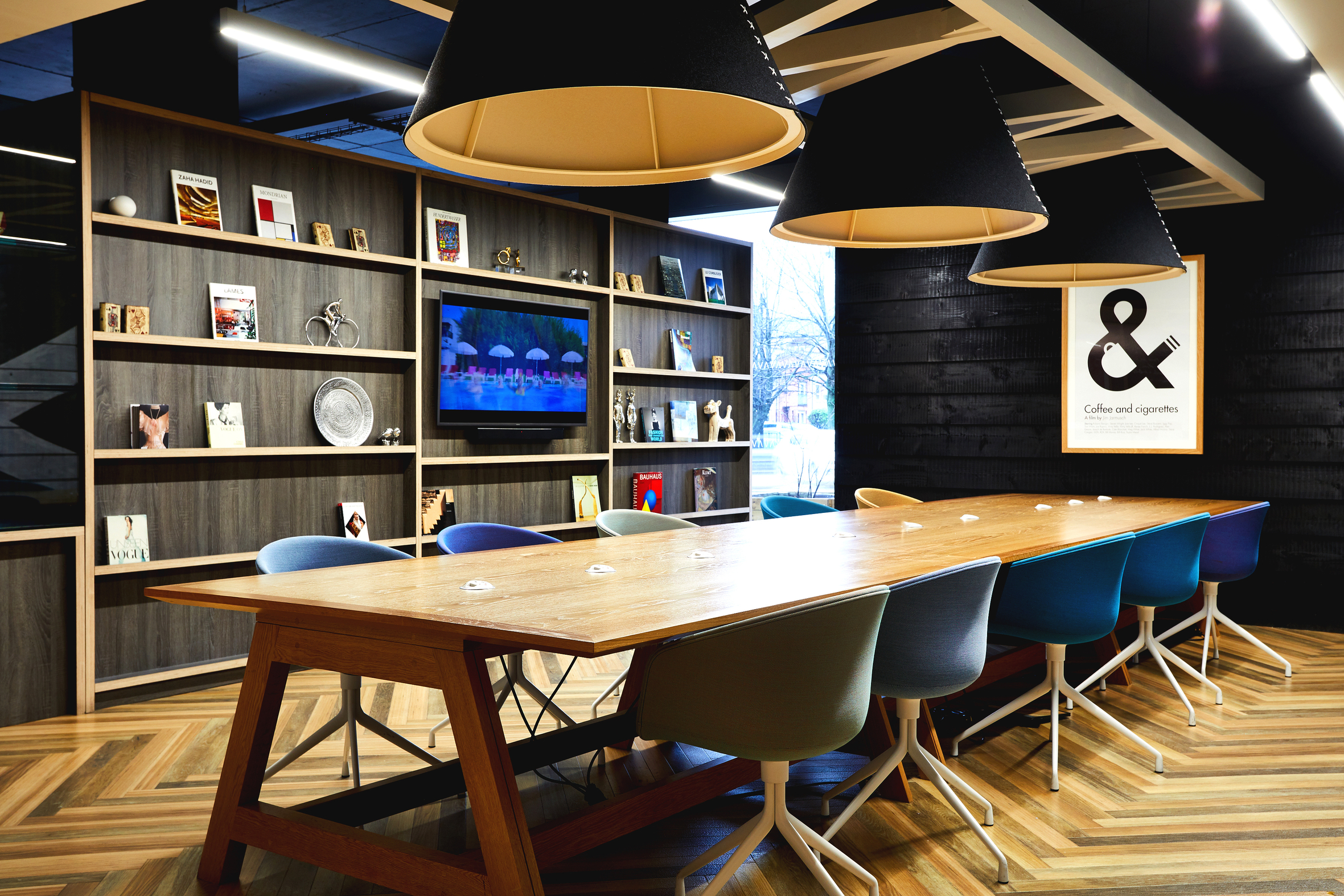 A modern conference room with a long wooden table surrounded by colorful chairs. Shelves with books and decor line the back wall. Large, black pendant lights hang from the ceiling, and a framed print is displayed on the wall.