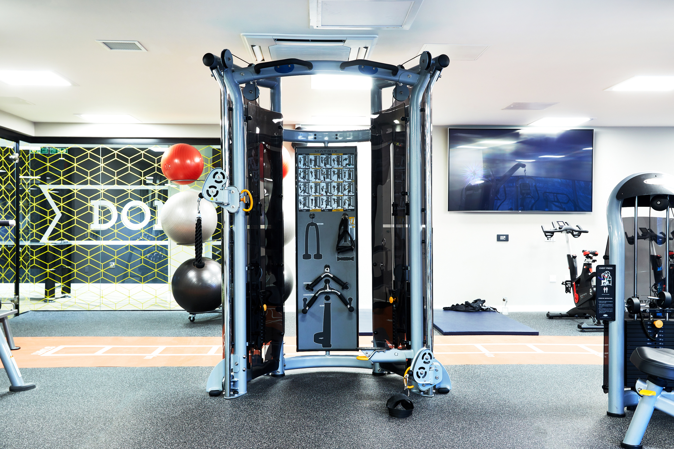 A modern gym with a functional trainer machine in the center. Exercise balls are stacked against a wall with the word "DOIT." There is a large screen on the wall and various gym equipment in the background.