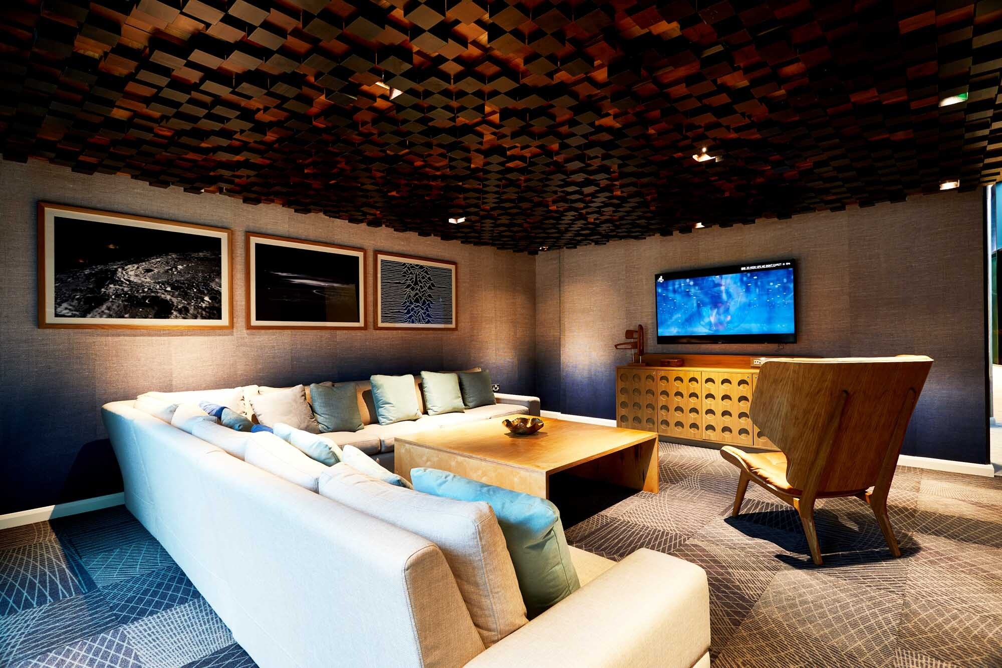 A cozy living room with a white sectional sofa and wooden armchair facing a mounted TV. The ceiling features a wooden block design, complemented by circular framed artworks on the wall. A large square wooden coffee table sits atop the patterned carpet flooring.