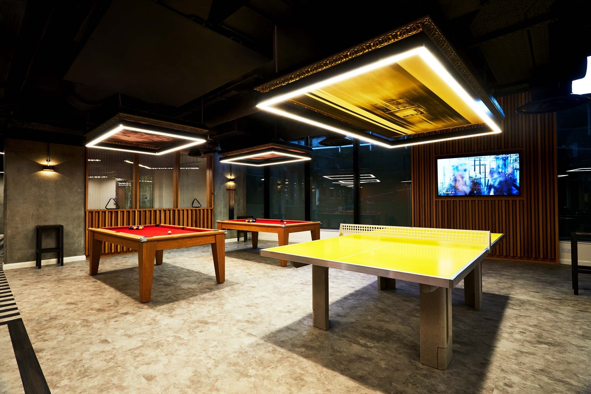 A modern game room features a yellow ping pong table and two red pool tables centered under square illuminated ceiling panels, reflecting the tables' colors. A wall-mounted TV is on the right, and the floor has a marble pattern. Warm lighting creates a cozy atmosphere.
