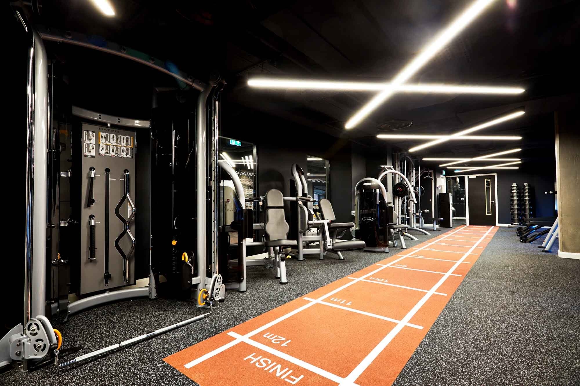 A modern gym boasts sleek exercise equipment, including weight machines and benches. A marked orange running track curves in a circle along the floor, leading to a mirrored wall. Overhead, bright strip lights create an X pattern, illuminating the square space beautifully.