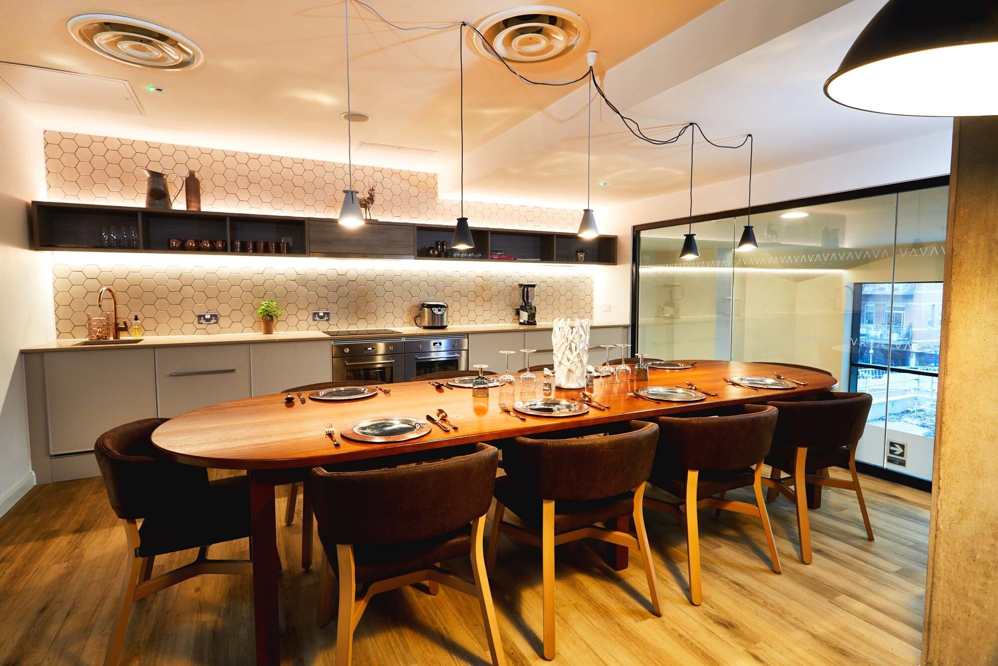 A modern dining room features a long wooden table set for eight, surrounded by dark chairs. Overhead circle pendant lights hang from the ceiling. The room boasts warm lighting, wooden flooring, and a sleek kitchen area with hexagonal backsplash tiles.