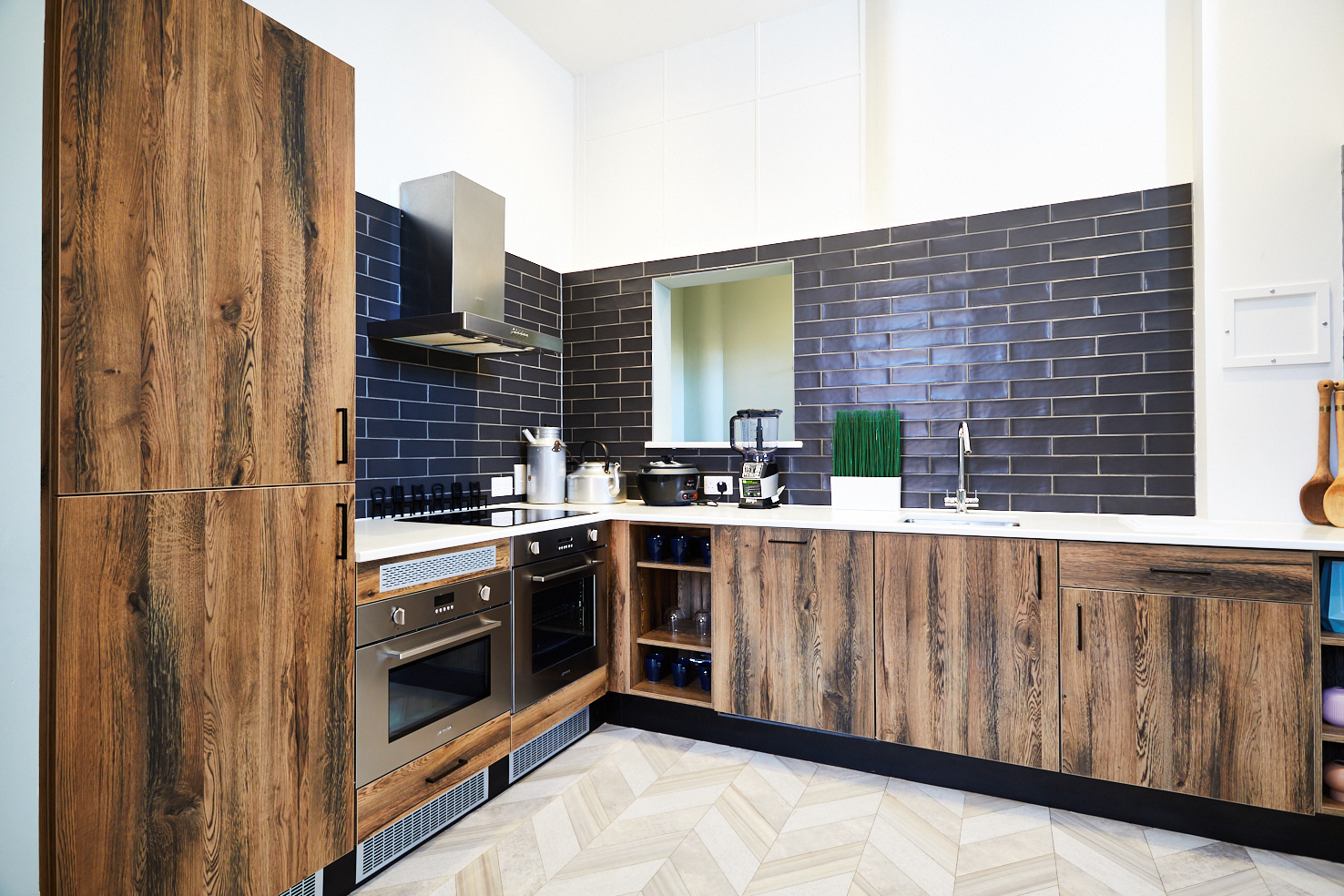 A modern kitchen on Lawrence Street features wooden cabinets and a stainless steel oven with a range hood. The dark subway tile backsplash contrasts beautifully with the light herringbone pattern floor, while small appliances and a plant add charm to the countertop.