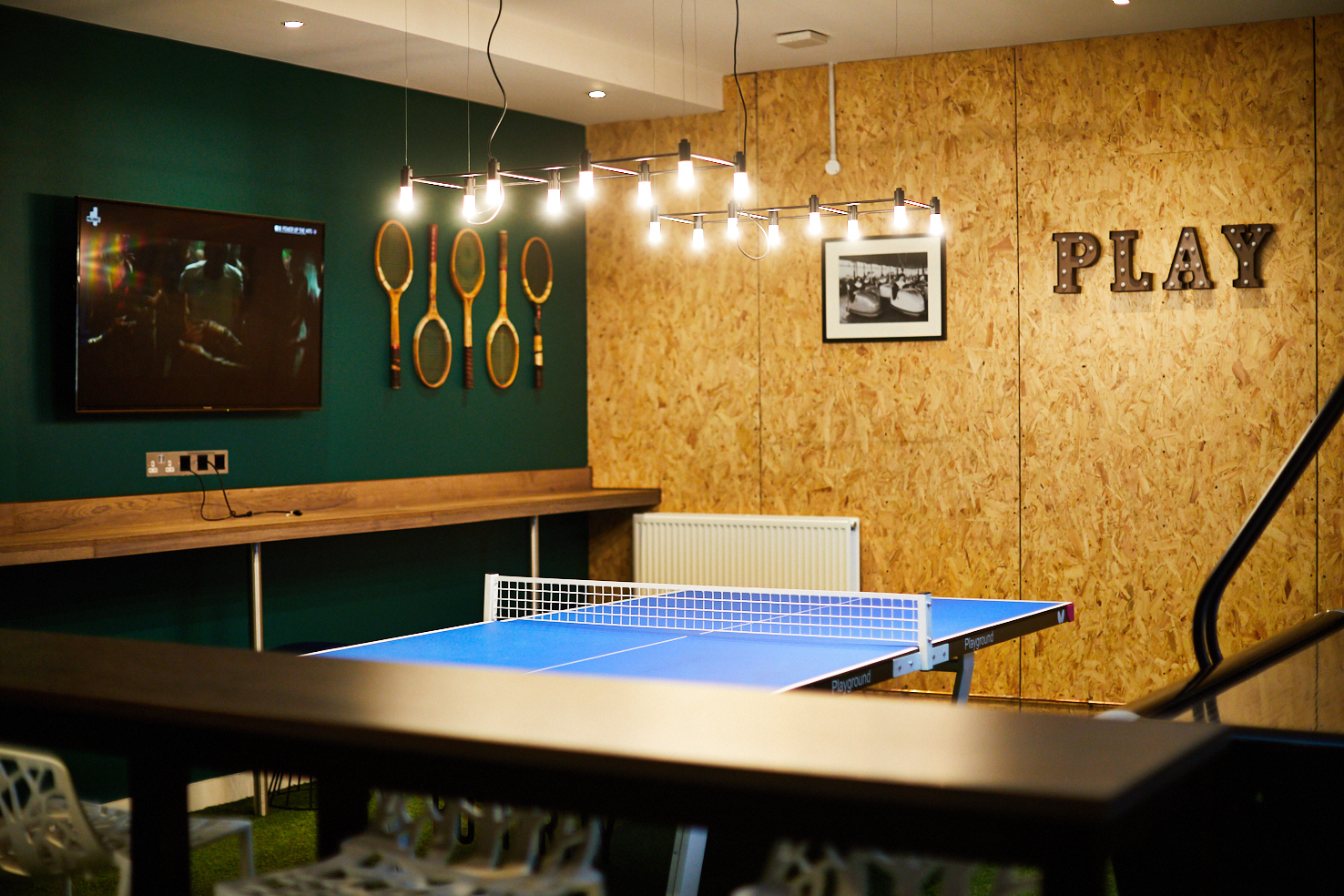 A modern game room on Lawrence Street features a blue ping pong table, mounted paddles on a wooden wall, and the word "PLAY" in large letters. A lit hanging light fixture and a wall-mounted TV screen displaying a program complete the vibrant space.