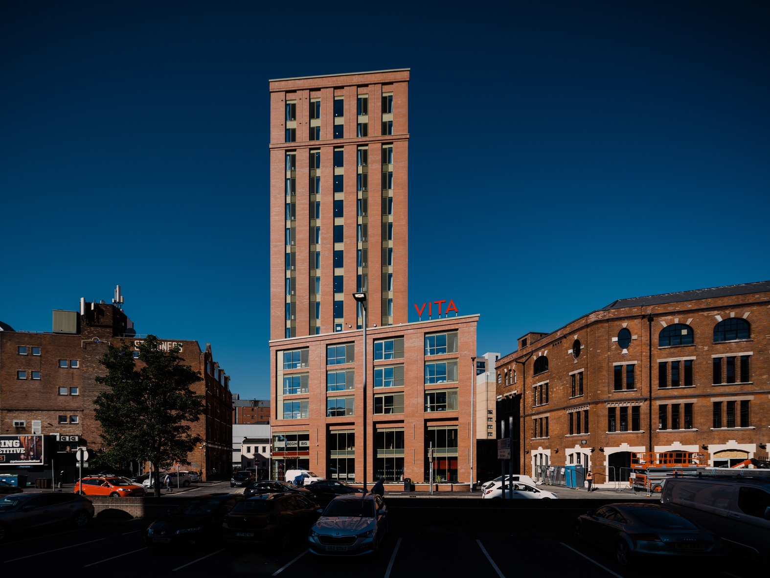 Tall red brick building with "VITA" signage, surrounded by lower brick buildings under a clear blue sky. A parking lot with cars is in the foreground.