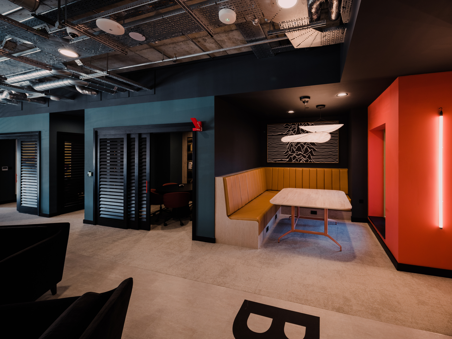 A modern office space with a cozy seating area featuring a yellow booth and a white table. The walls are dark with wooden accents, and a neon light is on the wall. Exposed ceiling ducts add an industrial feel.