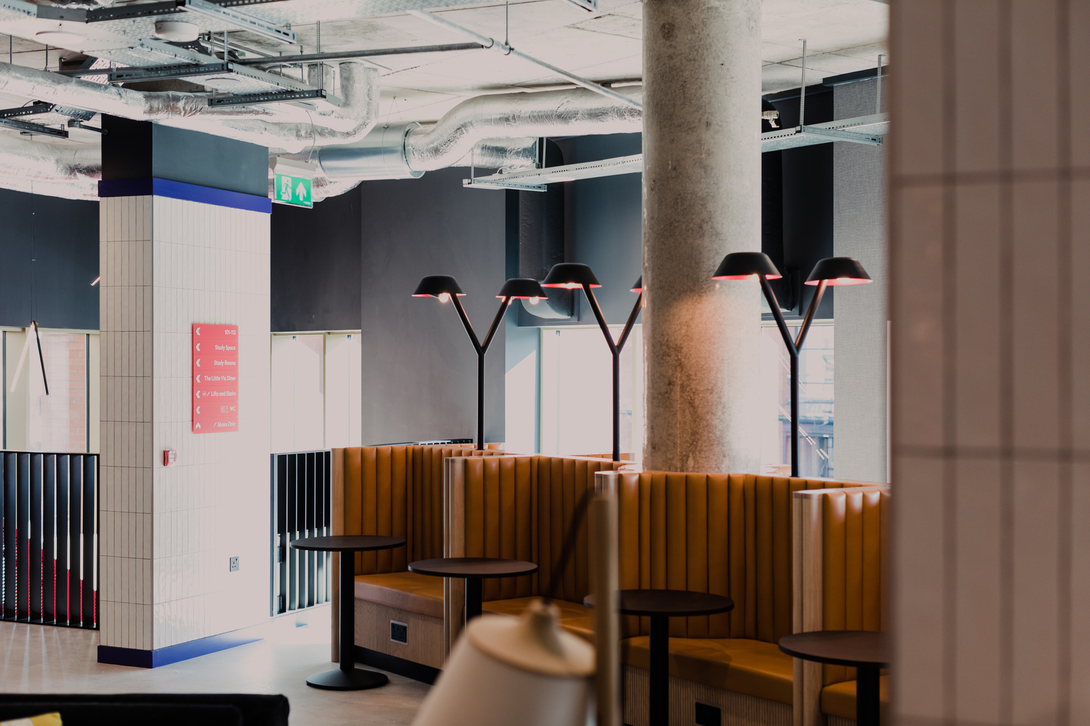 A modern cafe interior with mustard-colored booth seating against a wall. Black round tables sit in front of the booths. Overhead, black lamps emit warm light. The ceiling exposes industrial ducts. A red sign is visible on a tiled column.