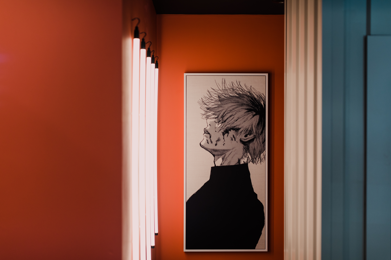 A framed artwork of a person with spiky hair and closed eyes, wearing a high-collared outfit, hangs on a vibrant orange wall. Vertical lights line the left side, creating a modern and artistic ambiance.