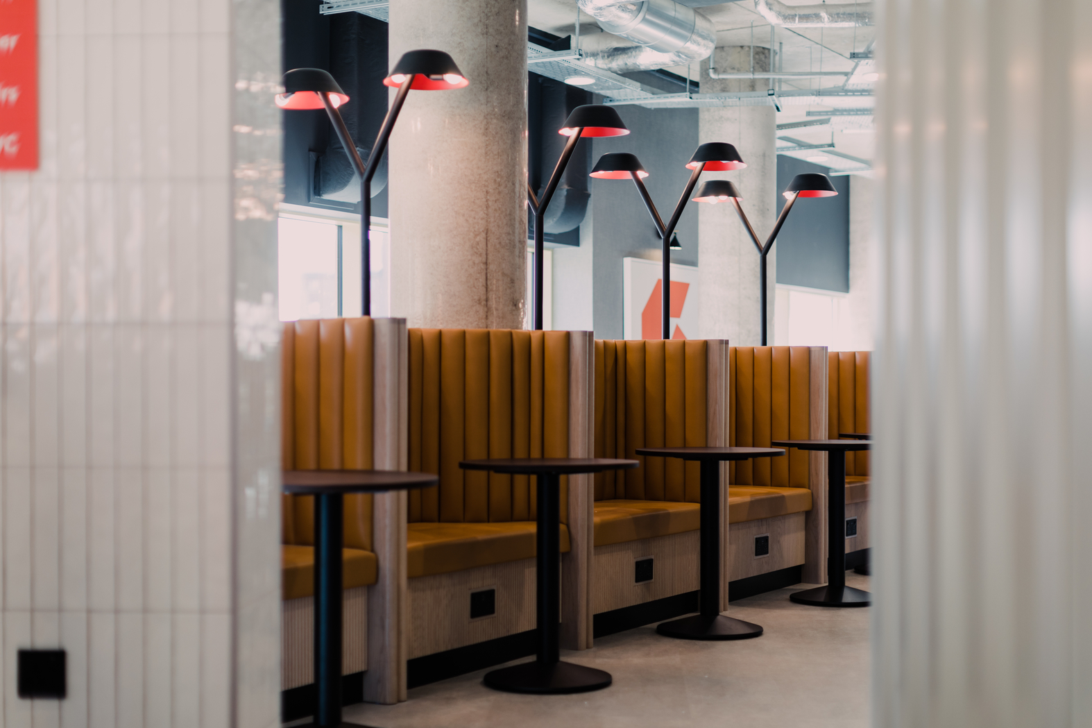 A modern interior with several wooden booths featuring mustard yellow cushioned seating. Each booth has a round black table. Overhead, there are stylish black lamps with red shades. Industrial elements are visible in the background.