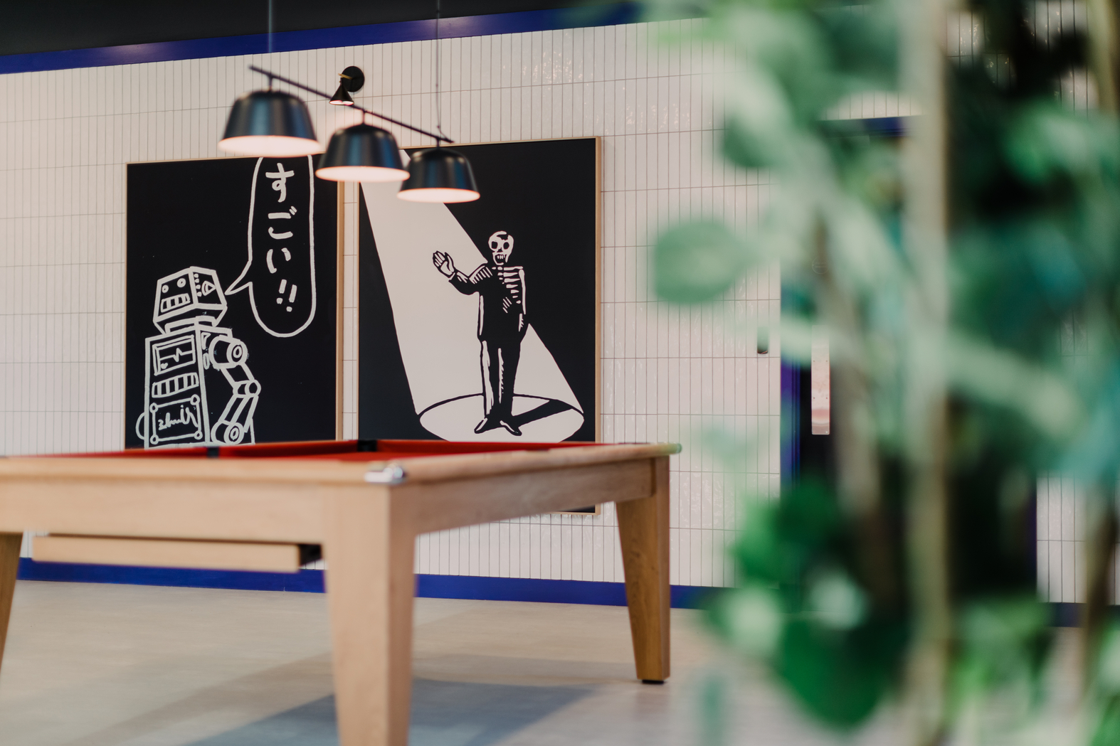 A billiard table indoors with modern art on the walls. One artwork shows a cartoonish robot with Japanese text, and the other features a person standing. A plant in the foreground slightly blurs the view.