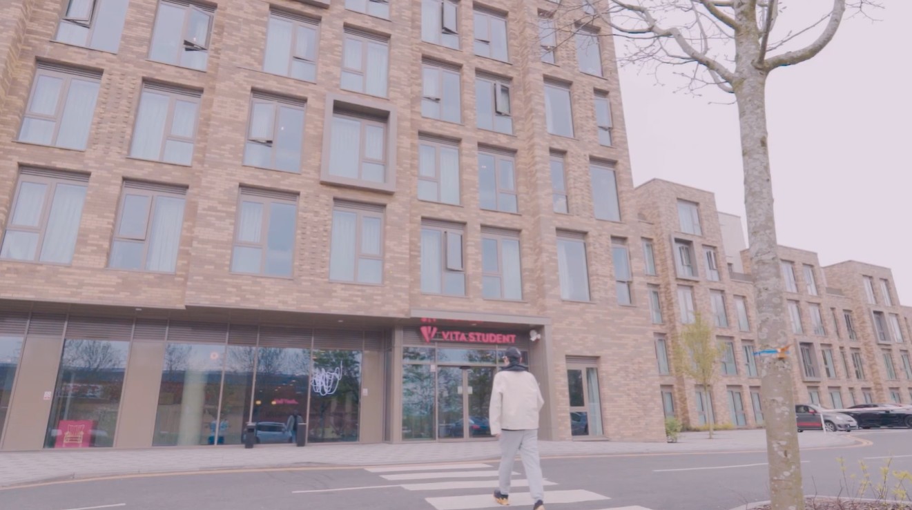 A person in a white jacket walks across a crosswalk toward a modern brick building with large windows and a "VITA STUDENT" sign, located near Warwick’s Cannon Park. A leafless tree stands on the right side of the image.