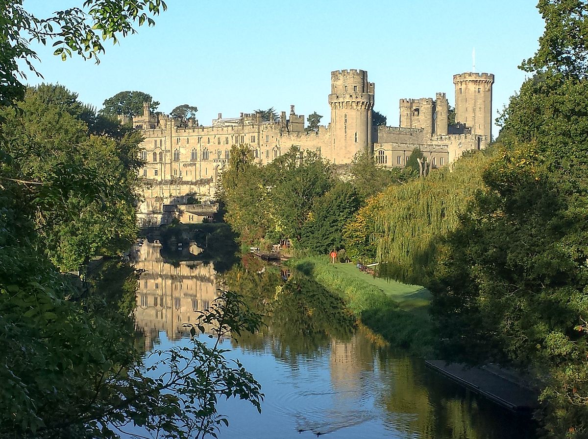 A scenic view of a large historic castle with multiple towers, surrounded by trees, reminiscent of the student accommodation in Coventry. It is reflected in a calm river in the foreground, with lush greenery on the riverbank under a clear sky.