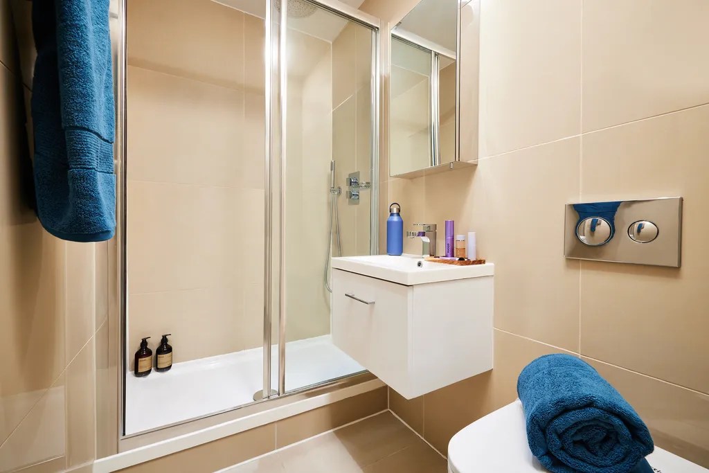 Modern bathroom with beige tiles, a glass shower enclosure, wall-mounted sink with toiletries, and a blue towel on the toilet seat. Another blue towel hangs on the wall. Two bottles sit in the shower corner.