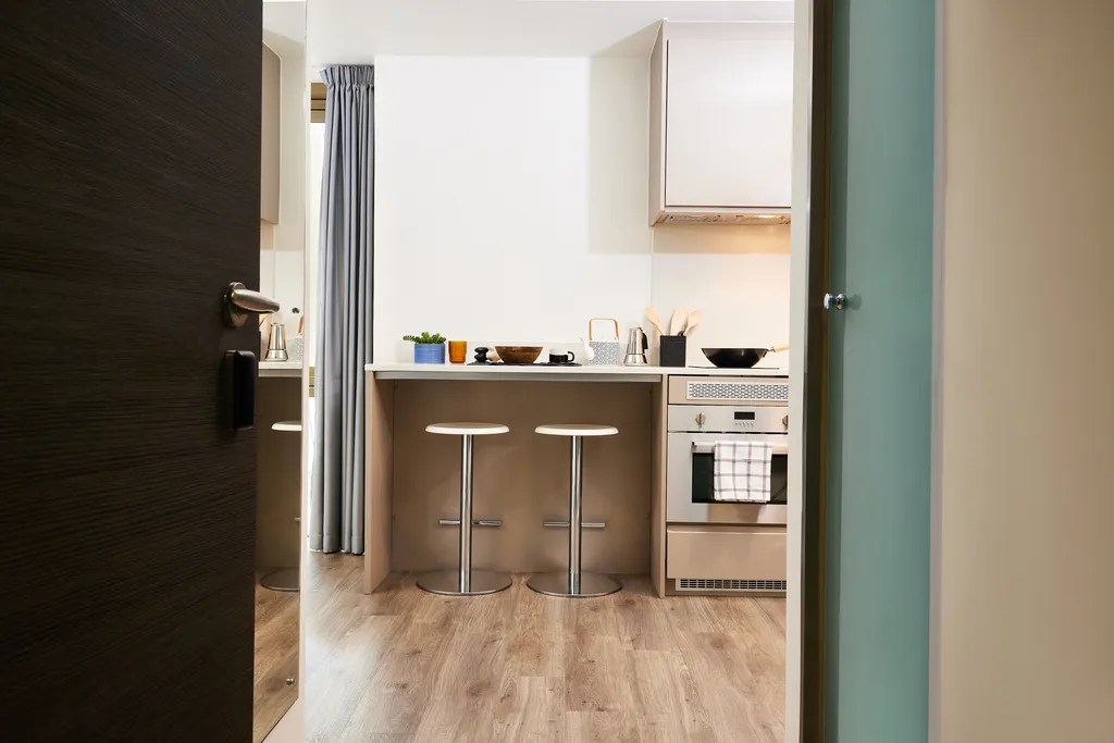 A modern kitchen with light wood flooring, a white countertop, two bar stools, an oven with a towel hanging, and various kitchen items neatly arranged. A door is partially open, revealing the space.