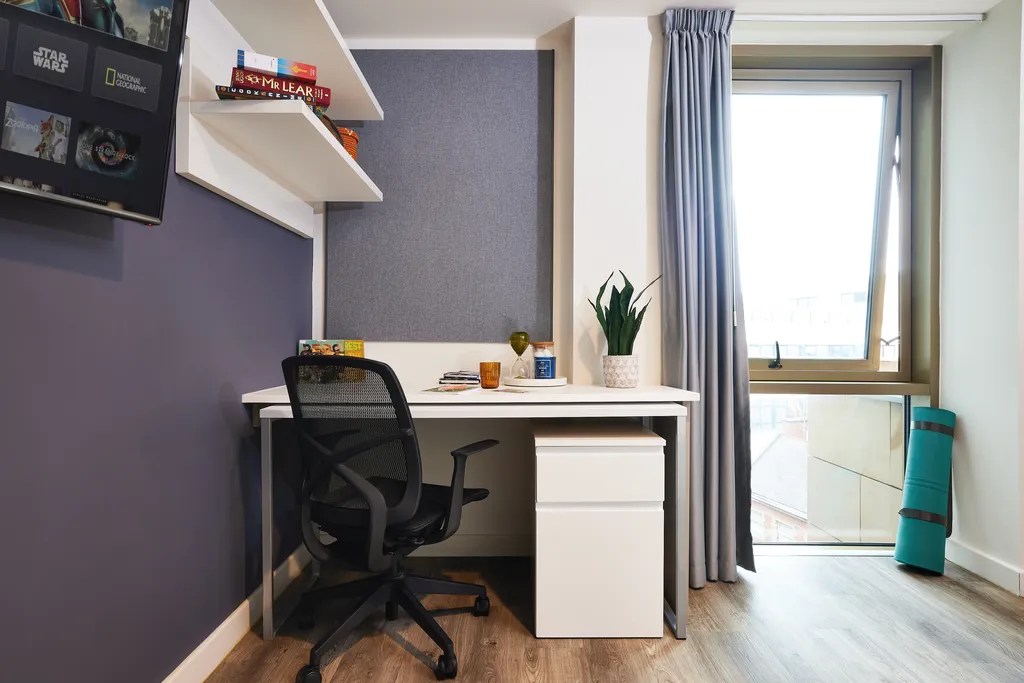 A modern home office with a white desk, black chair, potted plant, books, and cup. Shelves hold board games, and a window with curtains brings in natural light. A teal yoga mat leans against the wall on wooden flooring.