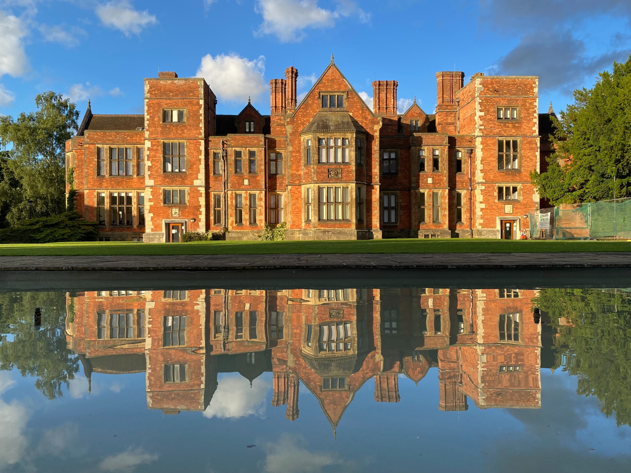 Historic brick mansion with numerous chimneys and windows, reflecting in a still body of water. The sky is blue with a few clouds, and trees frame the building on the right.